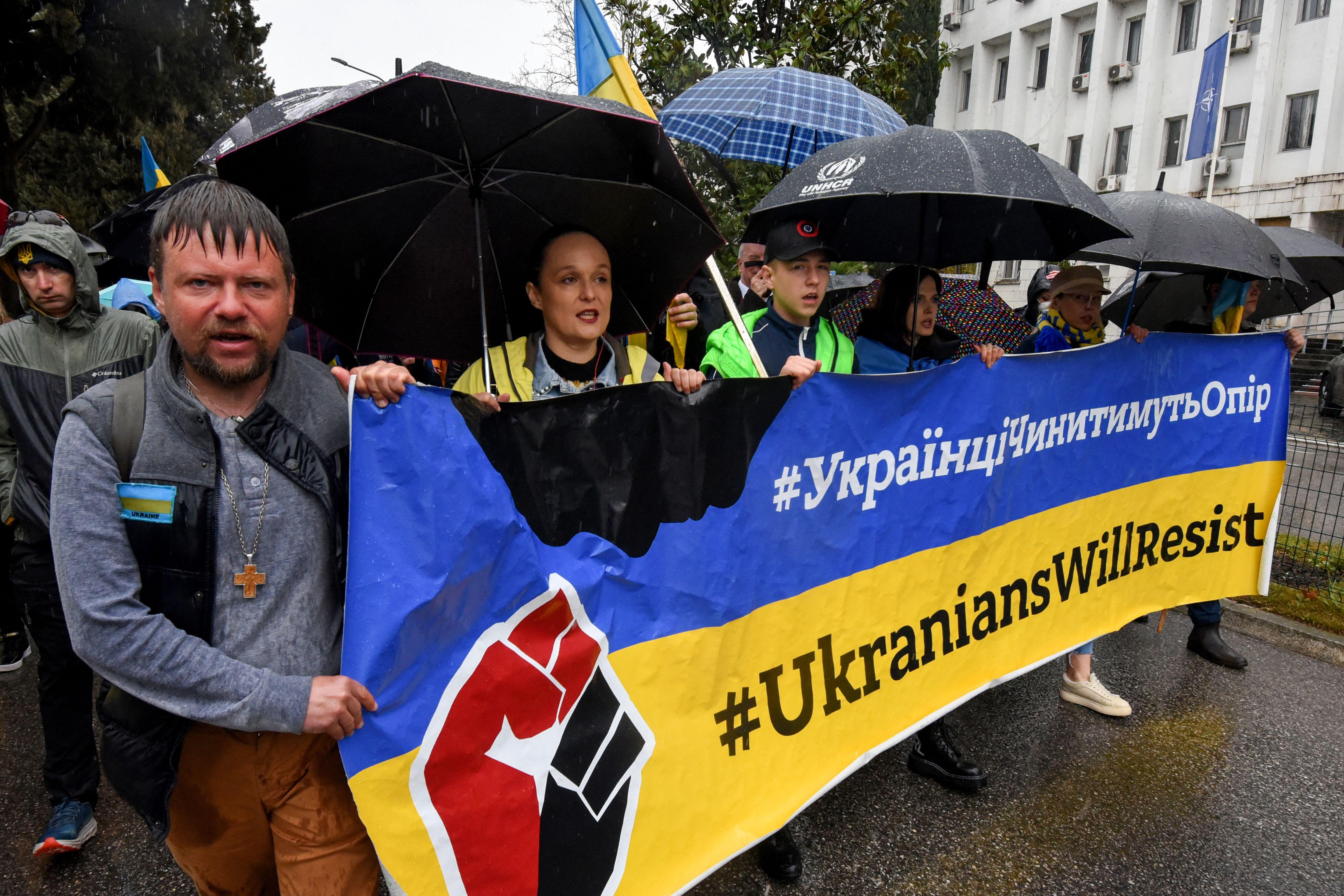 Several people hold a large, long sign that says #UkrainiansWillResist. Some are carrying umbrellas; a man on the end without an umbrella is soaking wet. 
