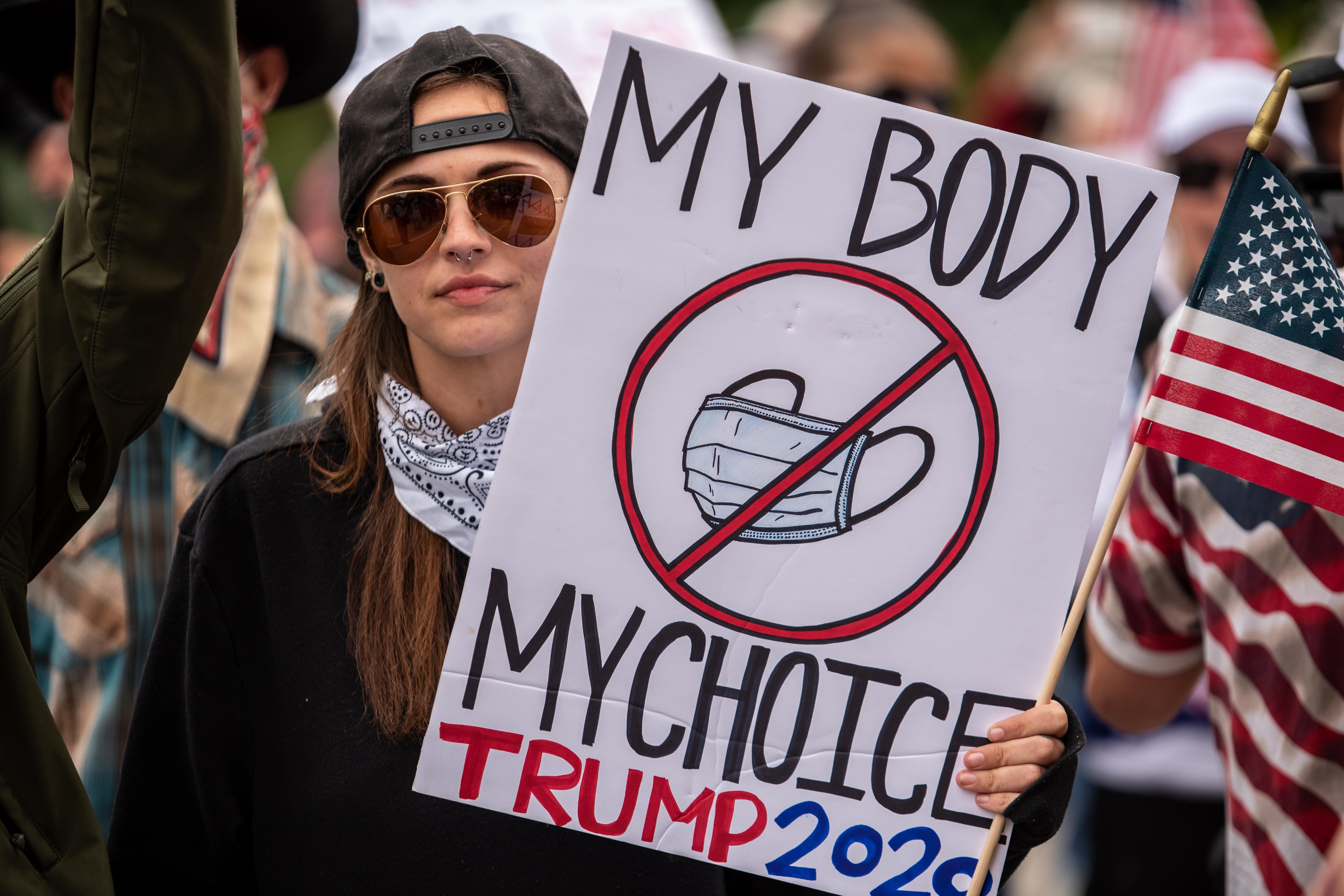 A woman holds up an American flag and an anti-mask-wearing sign that says "My Body My Choice Trump 2020"