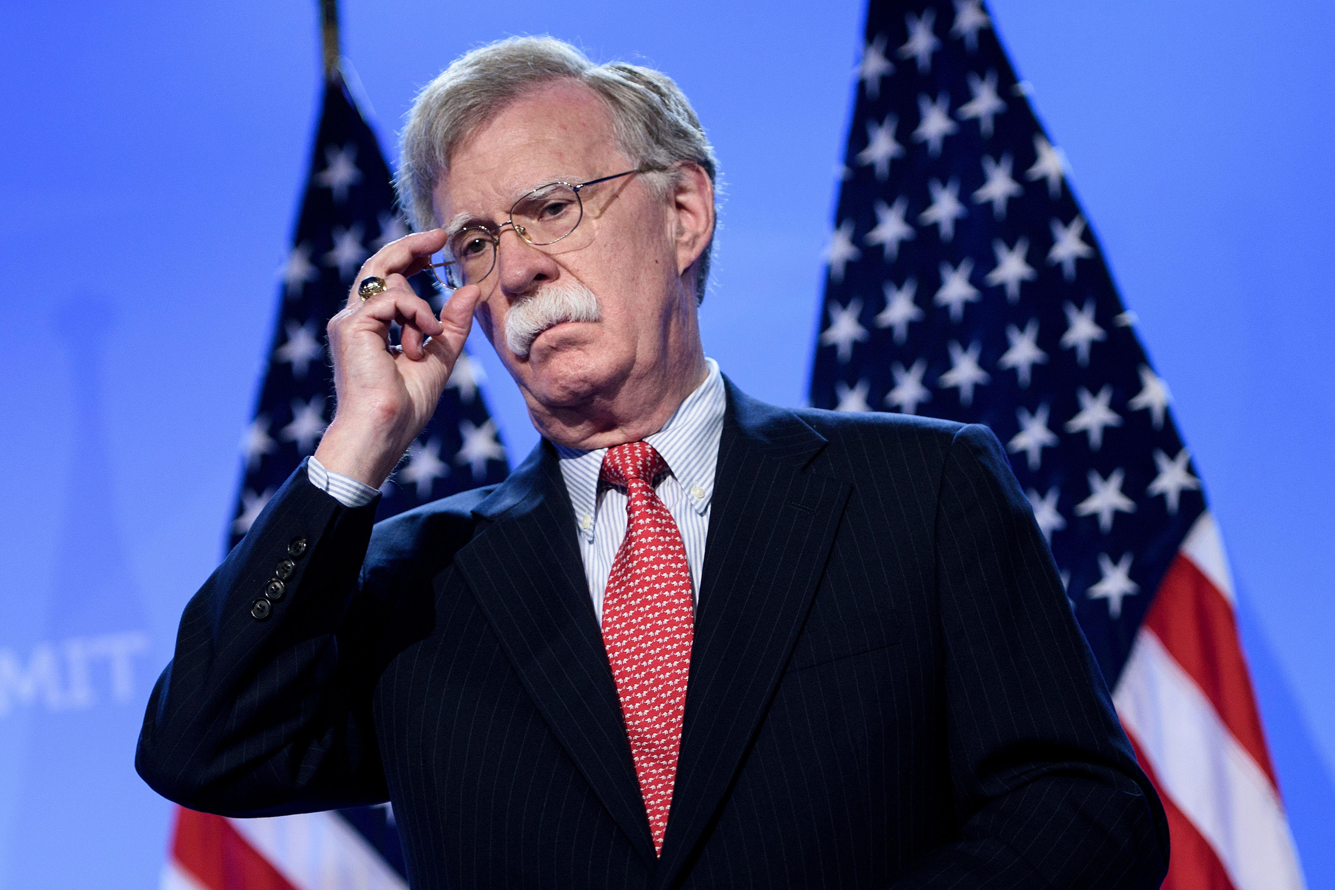 John Bolton adjusts his glasses in front of two American flags.