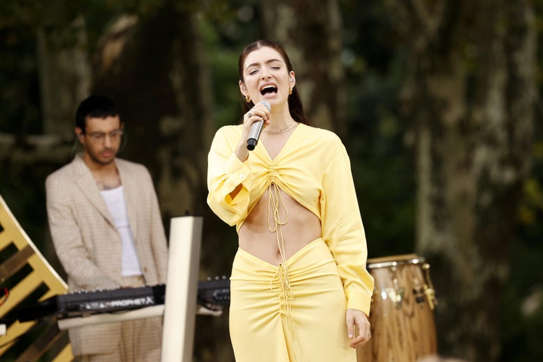 Lorde sings into a microphone while wearing a pale yellow crop top and long skirt.