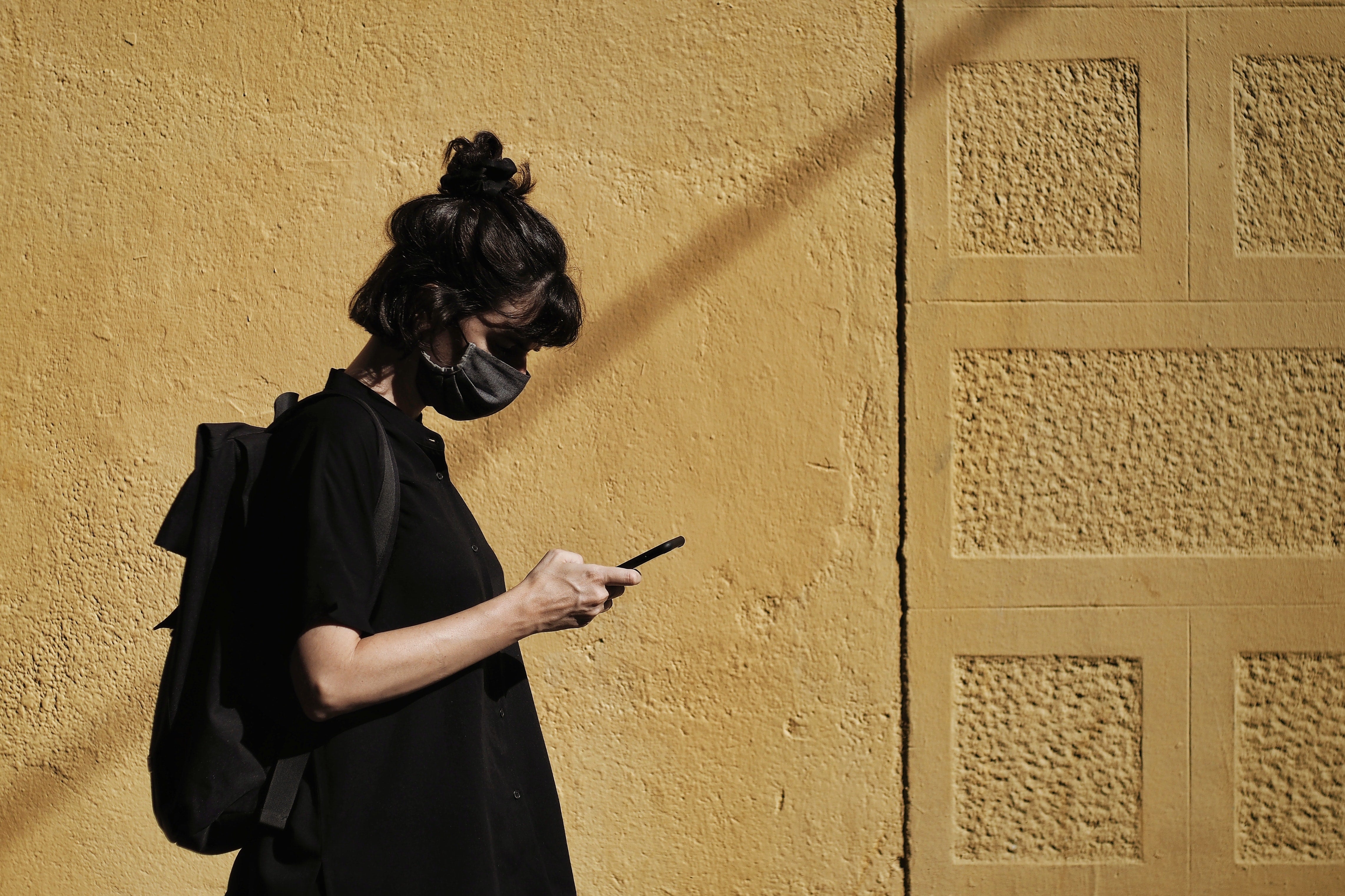 A woman wearing a black T-shirt and a black mask looking at her phone. She is standing in front of a yellow wall. 