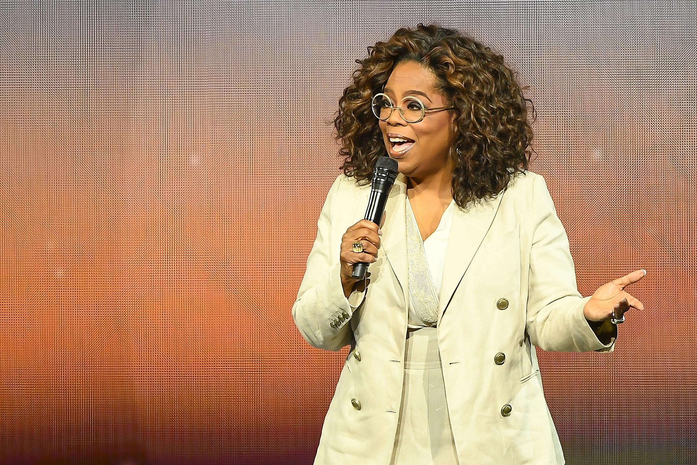 Oprah Winfrey dressed in beige blazer and round glasses talks into a microphone.