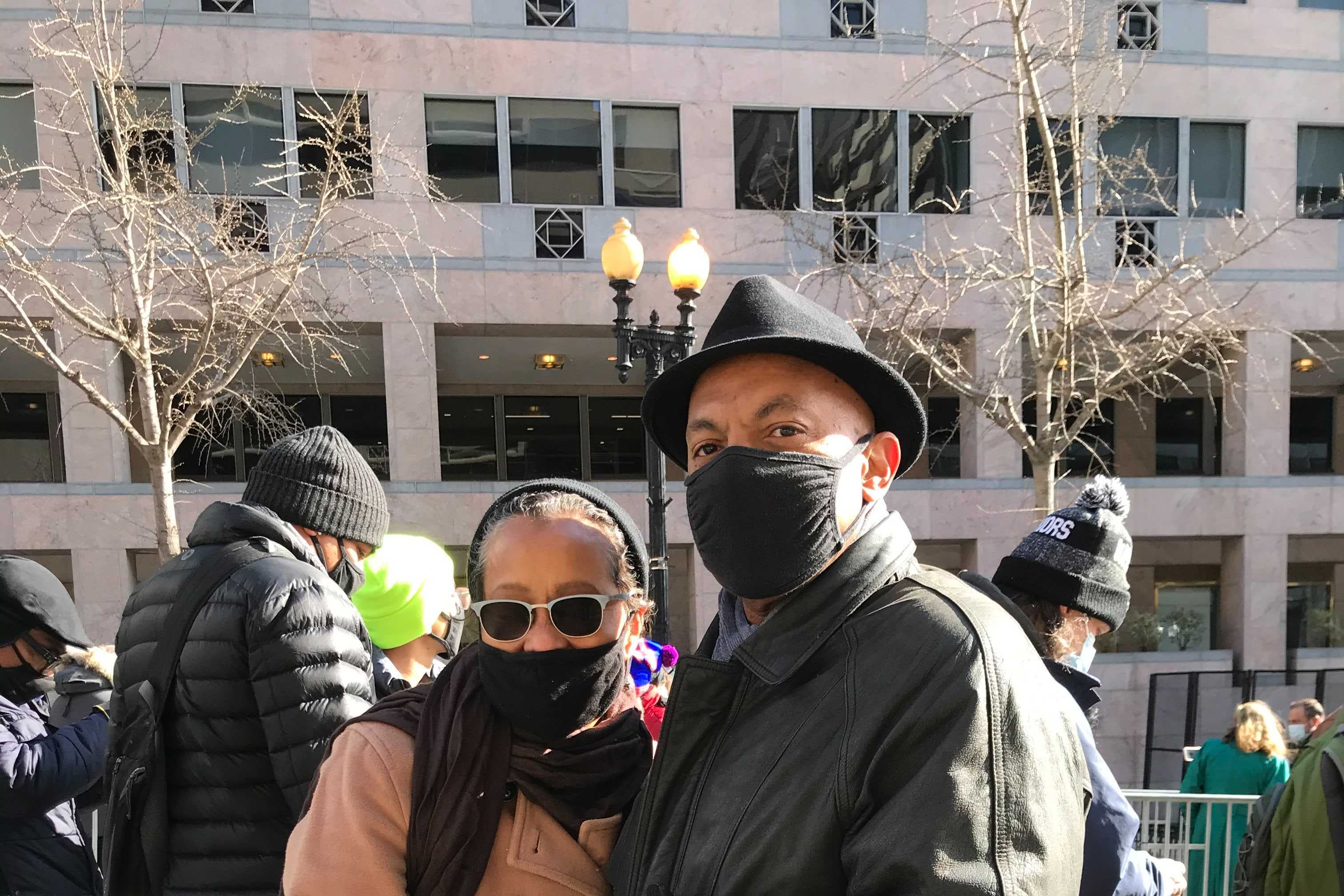 Two people wearing black masks stand in front of a metal barrier