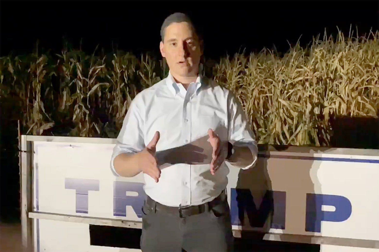 Still of Mandel in a white button-down shirt gesturing as he stands in front of a large TRUMP sign in a cornfield at night