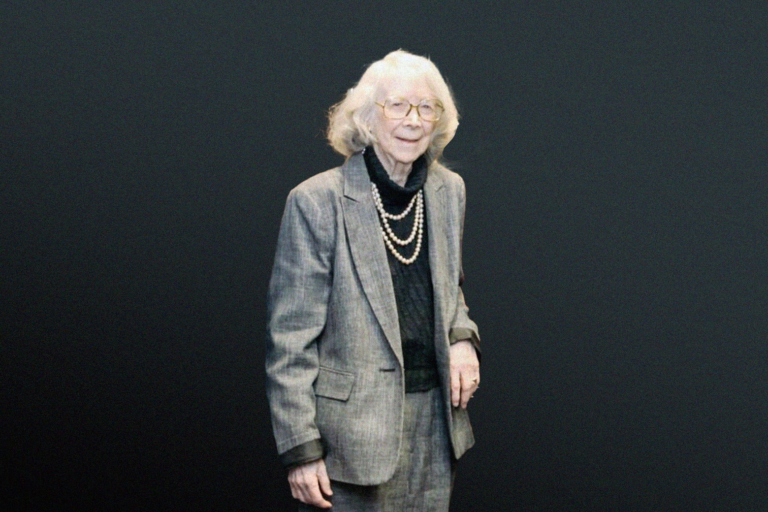 An elderly woman in a gray suit wearing large glasses, smiling.