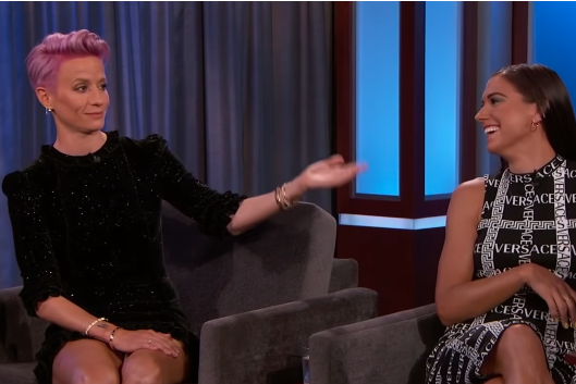 Megan Rapinoe gestures toward Alex Morgan, wearing a dress that says "Versace," as they both sit on a stage.