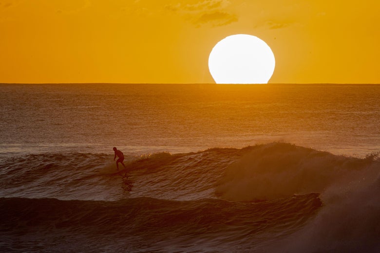 A surfer rides a wave as the sun sets on the horizon.