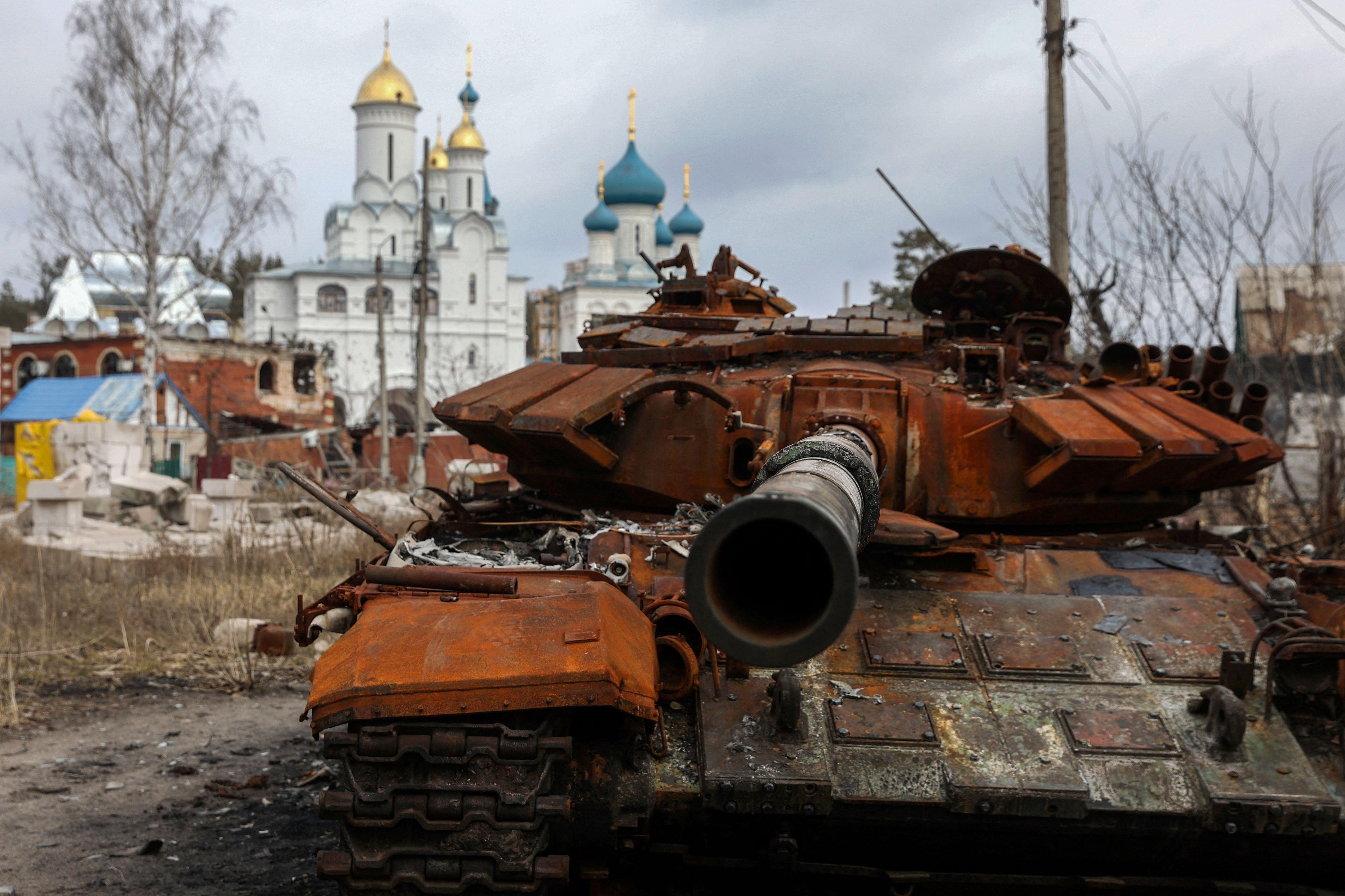 A destroyed Russian tank, rusted orange, sits abandoned. A church can be seen in the background.