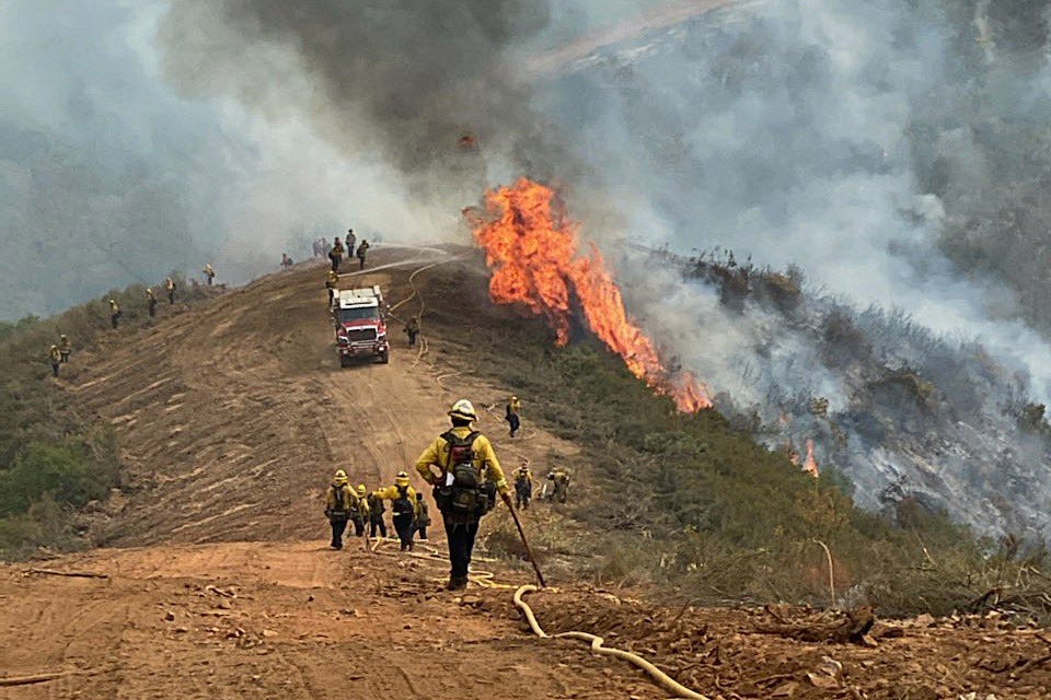 Firefighters standing near a fire, surrounded by smoke