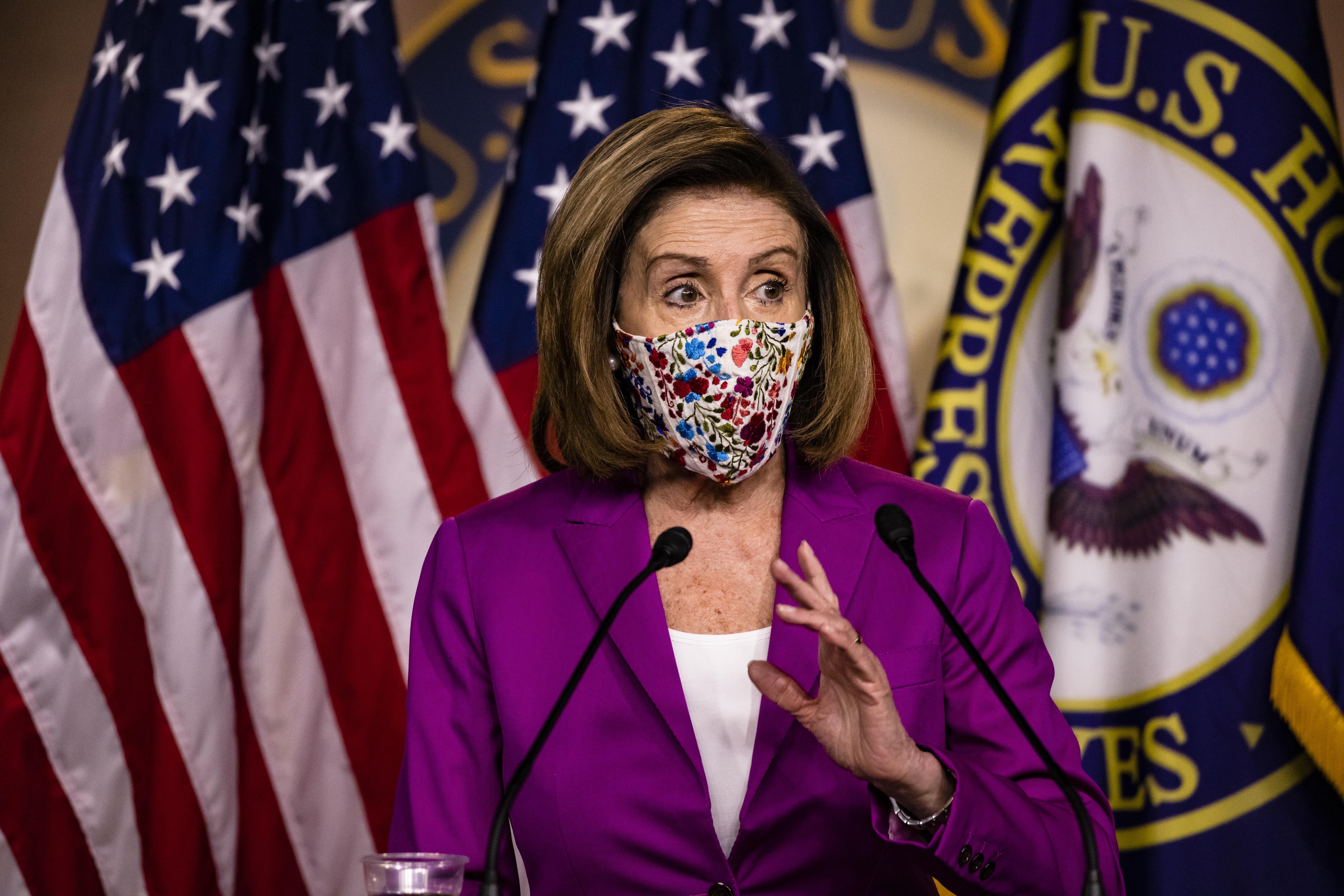 Pelosi, wearing a floral mask, speaks at a mic with American flags behind her
