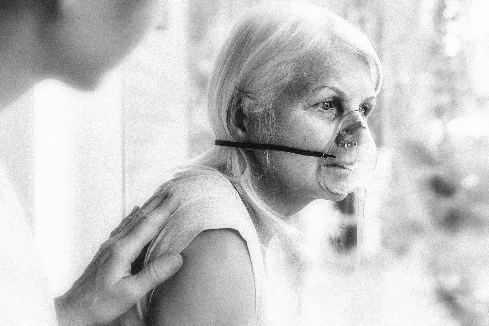 An older woman sits with an oxygen mask on her face while someone mostly out of frame puts a hand on her back.