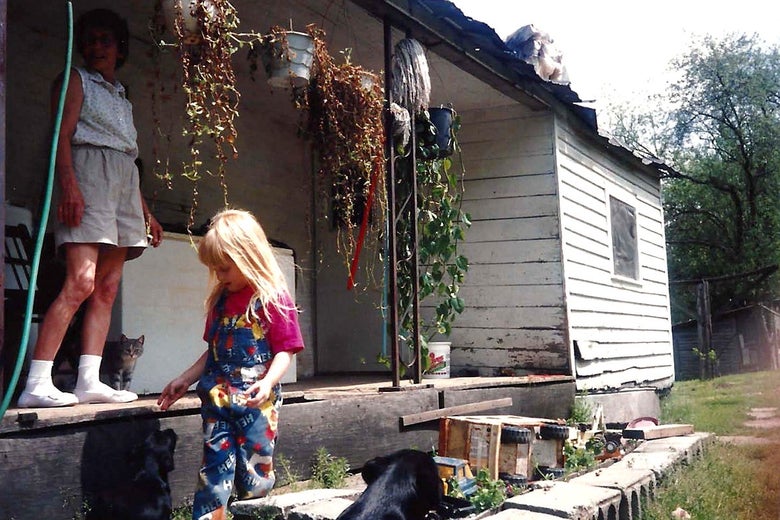 A blond child walks beside a porch with plants hanging from the roof. A woman wearing khaki shorts and white sneakers stands on the porch with a tabby cat poking out from behind her legs.