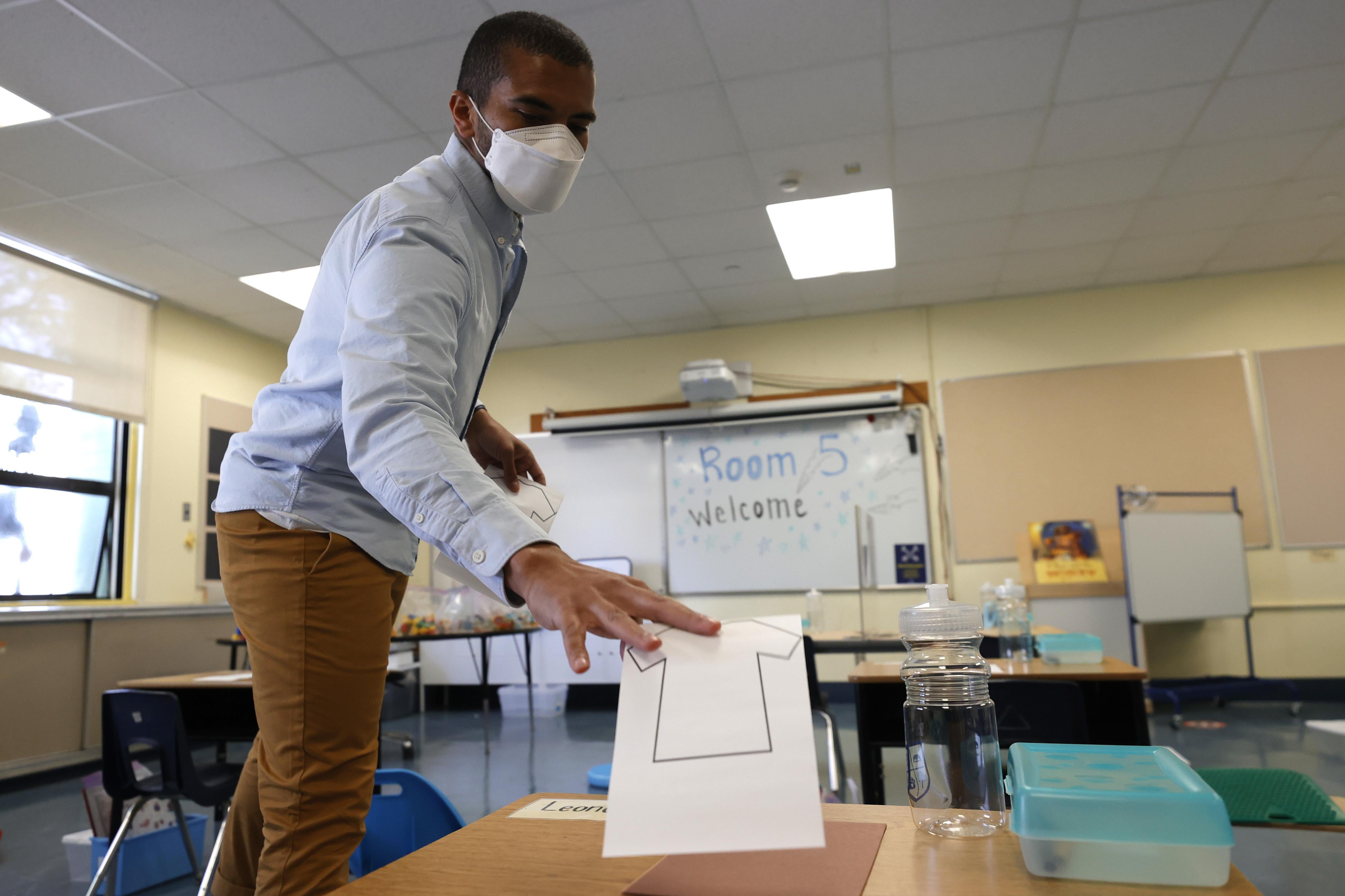 A Black man in a facemask puts a piece of paper on a desk in a classroom.