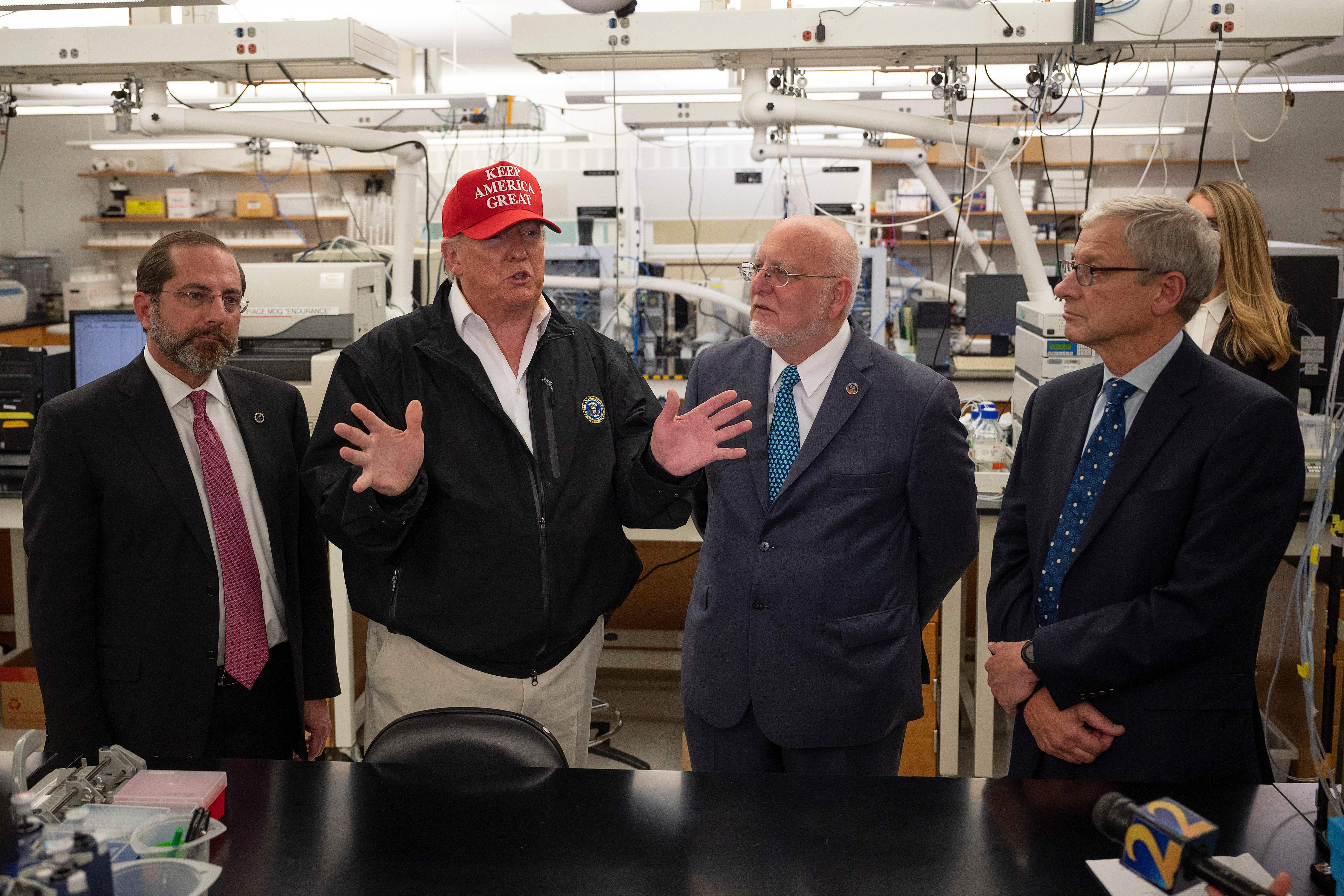 Azar, Trump, Redfield, and Monroe stand in a lab. Trump gestures with his hands and wears a MAGA hat.
