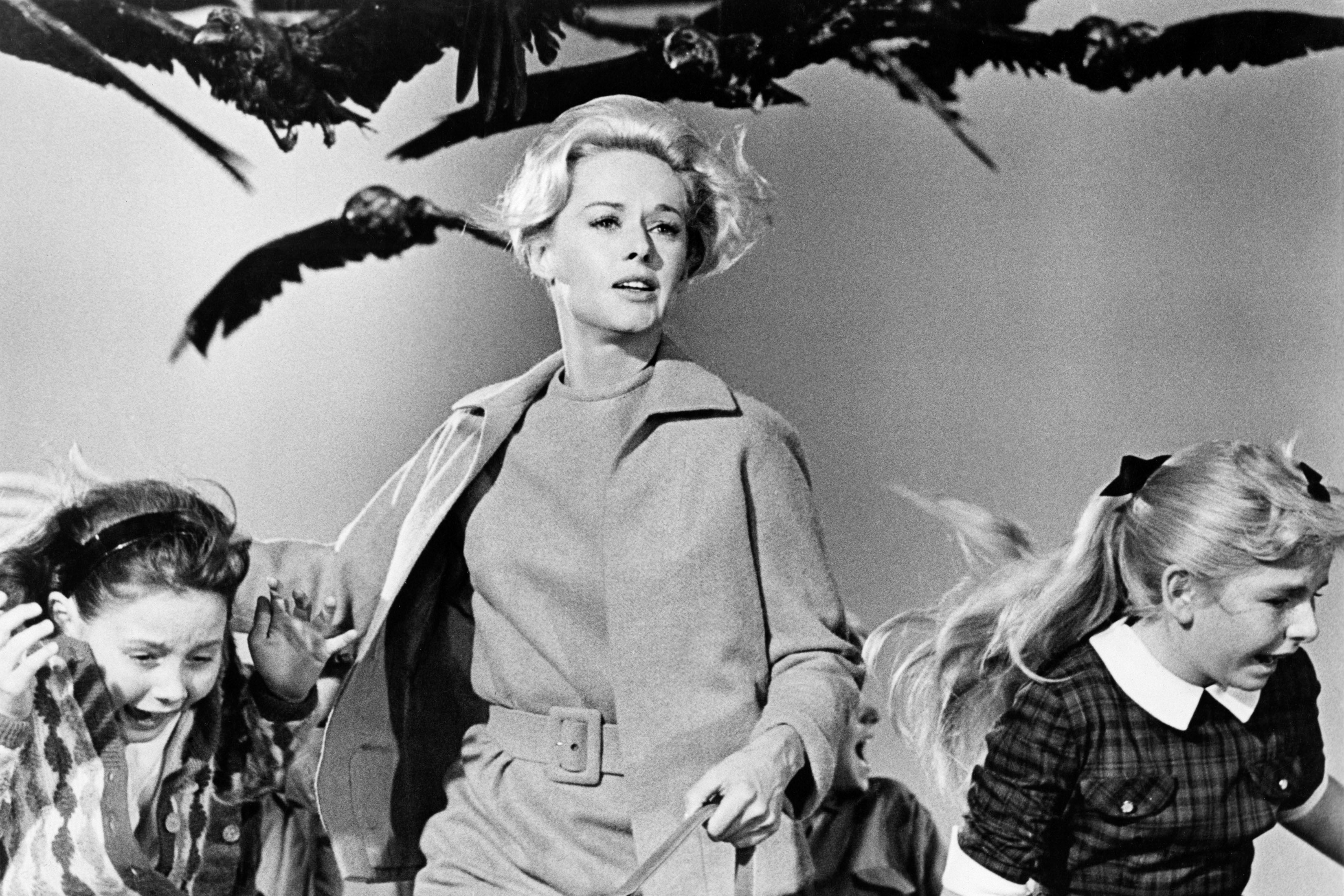 In black and white, an adult women runs along with two screaming girls as birds loom behind them. 