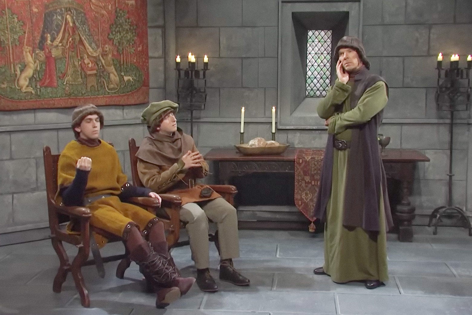 Benedict Cumberbatch standing in contemplation beside two cast members sitting in chairs, all three dressed in medieval garb, in a castle room