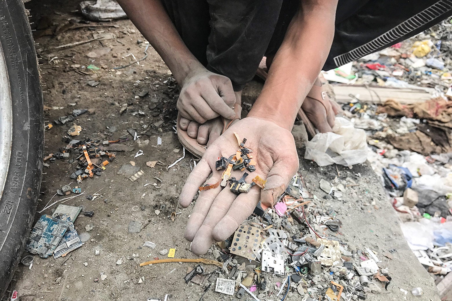 A boy, crouched, extends his hand, which is filled with small chips and other cellphone parts.