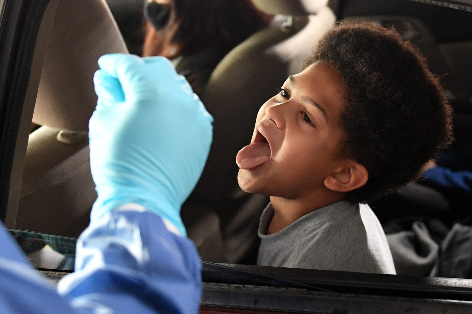 A boy sitting in the back seat of a car sticks out his tongue as a health care worker approaches.