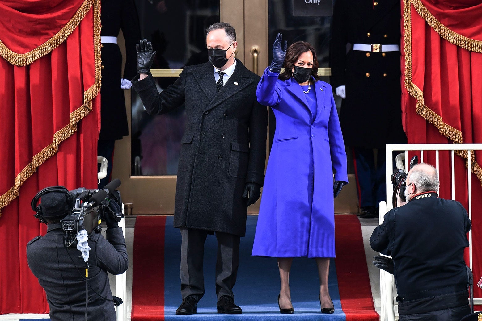 Doug Emhoff and Kamala Harris wave in front of the Capitol doors. Emhoff wears a dark suit, overcoat, and mask. Harris wears a purple coat with a purple dress underneath and a black mask.