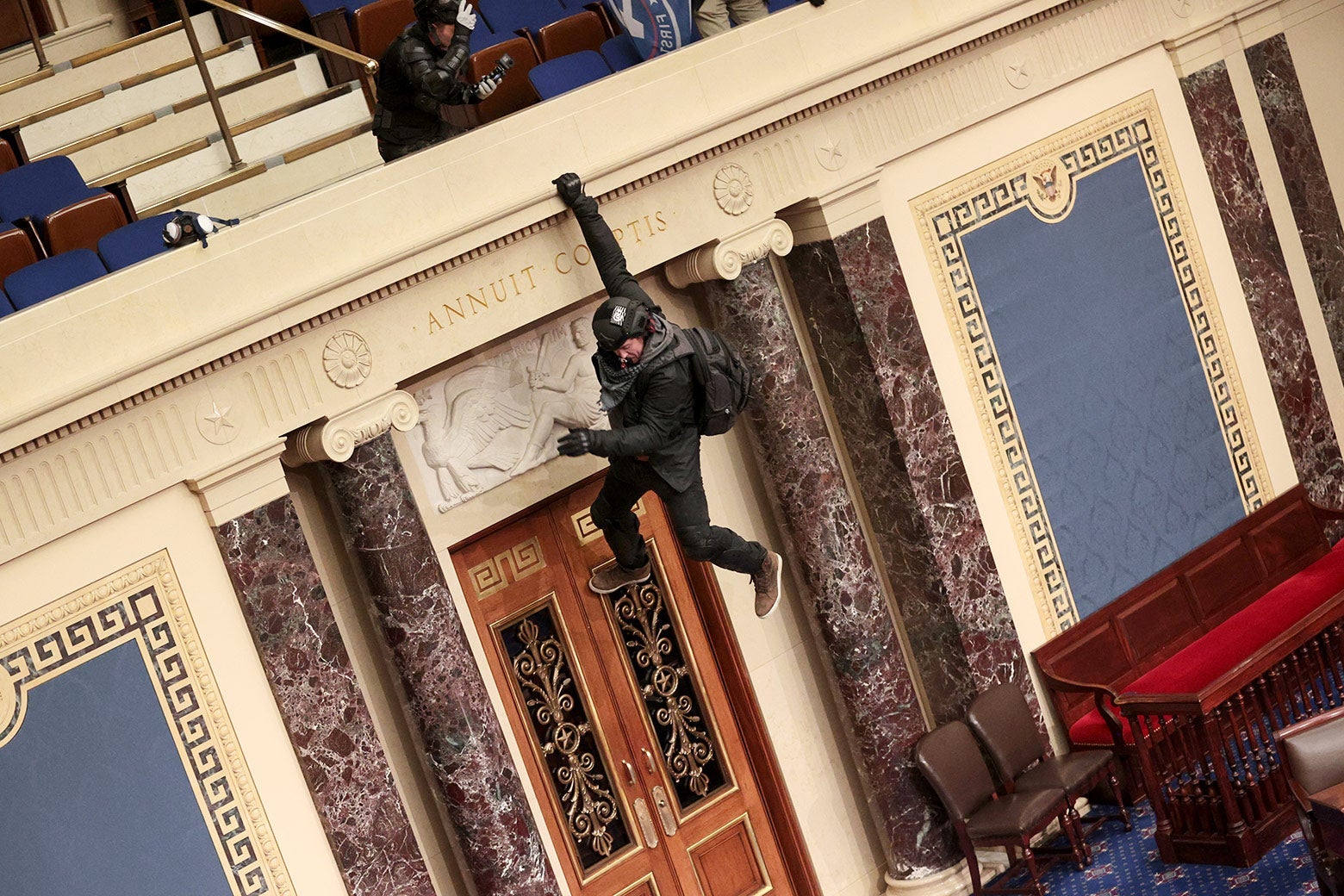A man with a backpack swings from the Senate chamber's balcony. He is hanging on with one hand.