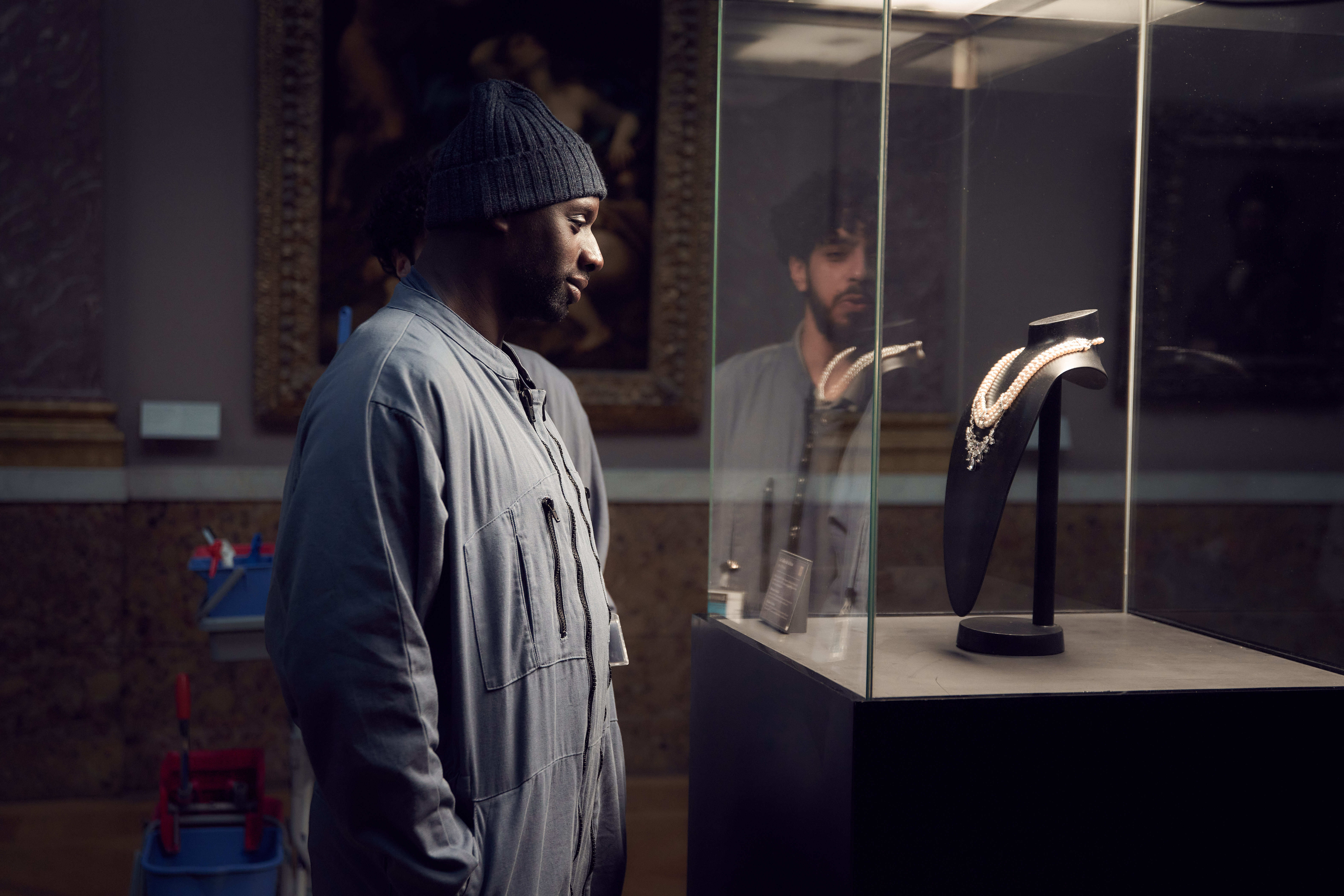Omar Sy stands in a dimly lit gallery, looking into a glass case containing a necklace.