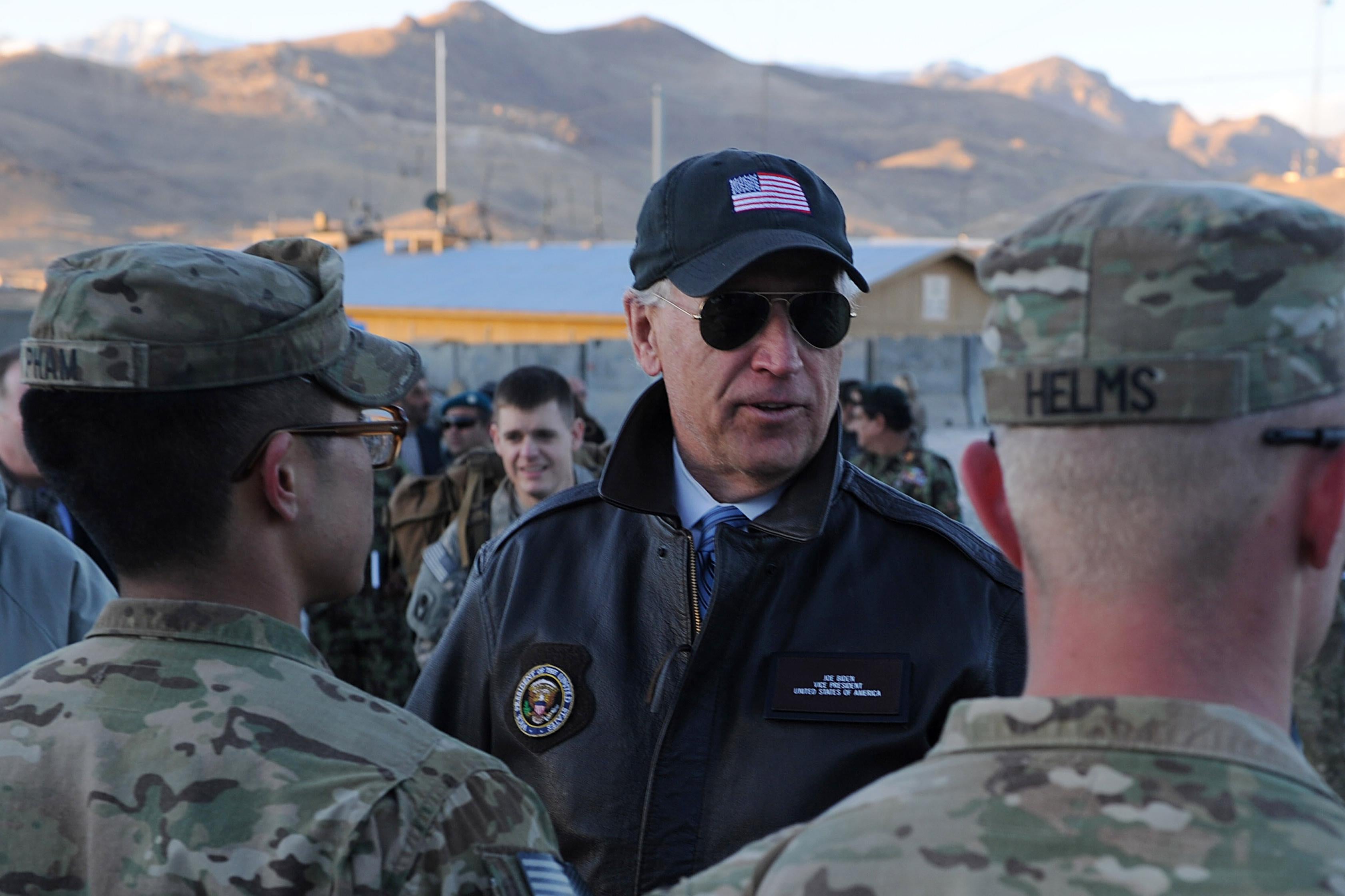 Biden in an American flag hat speaks to two soldiers with mountains and a helicopter in the background. 