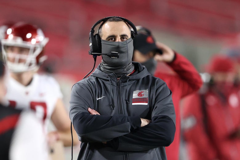Rolovich in headset and mask with his arms folded on the sidelines, looking upward with players behind him