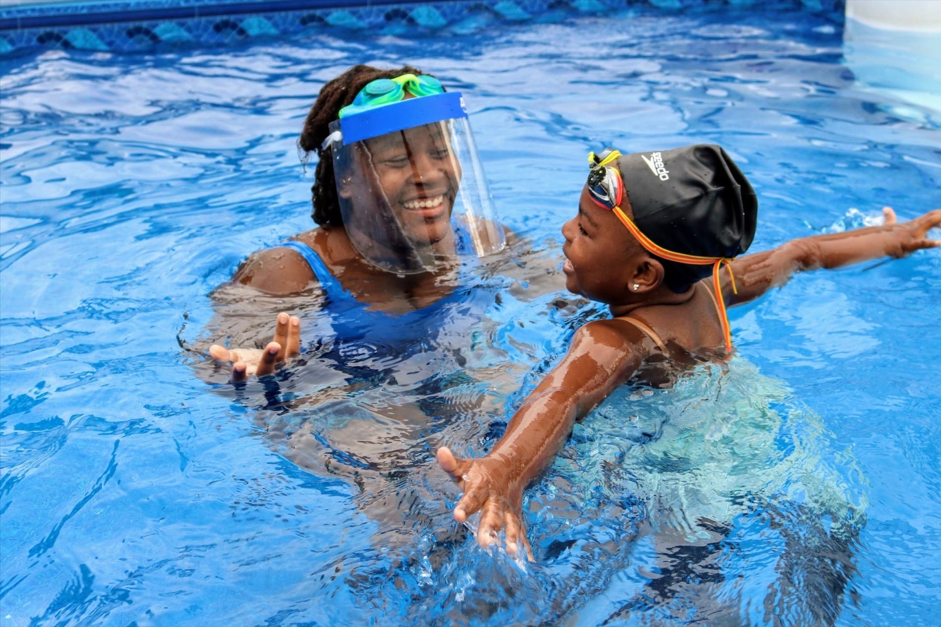 A woman wearing a face shield teaches a kid to swim in a pool