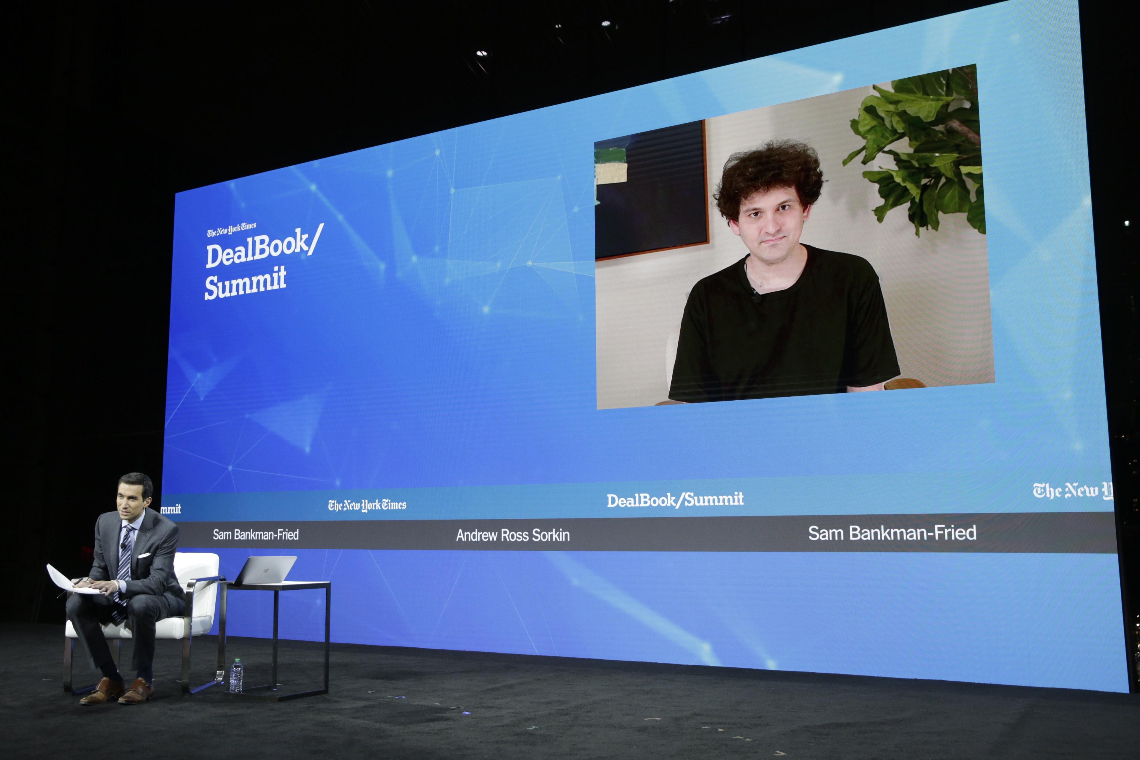 Andrew Ross Sorkin sitting in front of a giant DealBook-themed screen showing Sam Bankman-Fried on a video call.