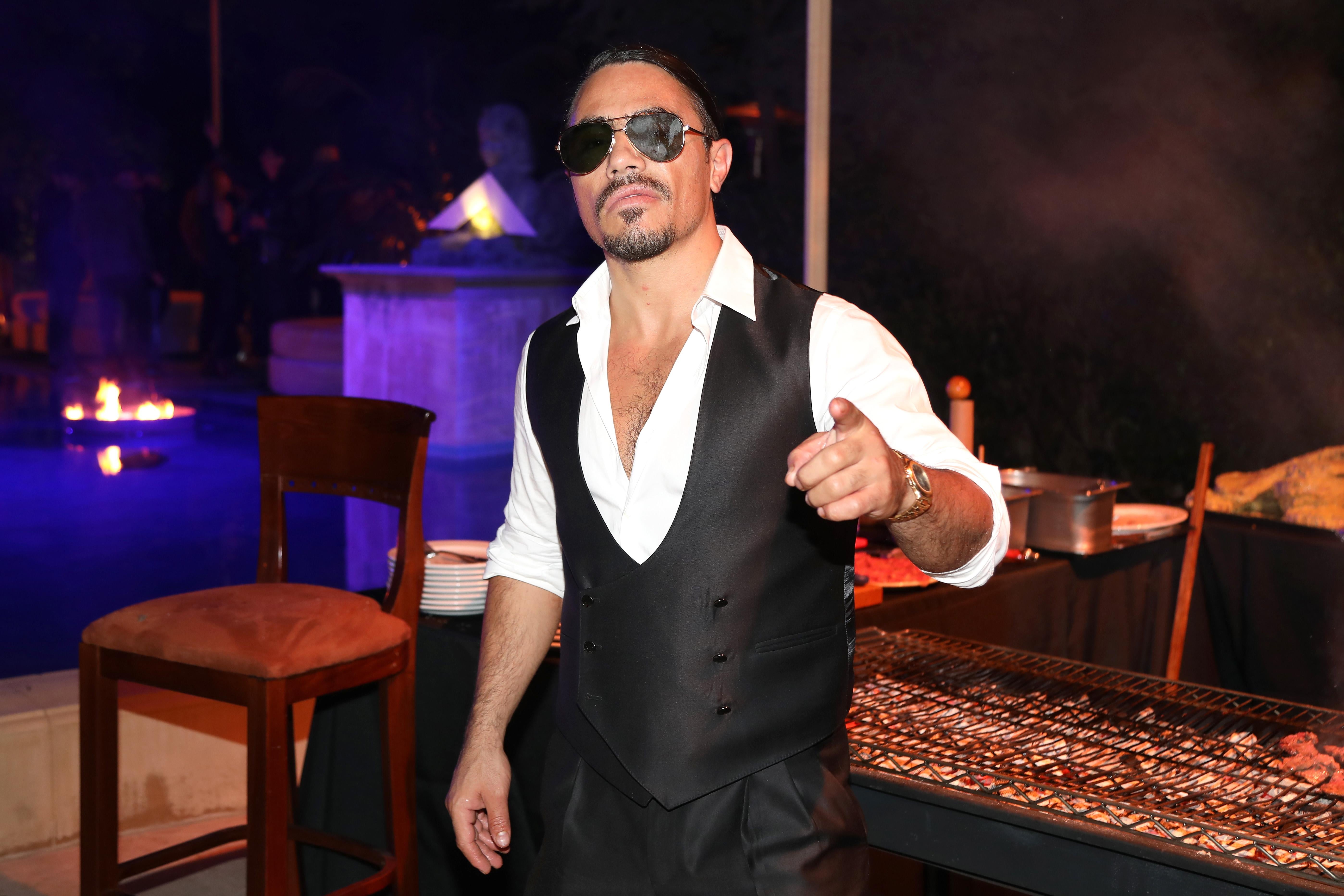 Nusret Gökçe, wearing a low-buttoned shirt and vest with sunglasses, points toward the camera while standing in front of a grill at a partylike setting.