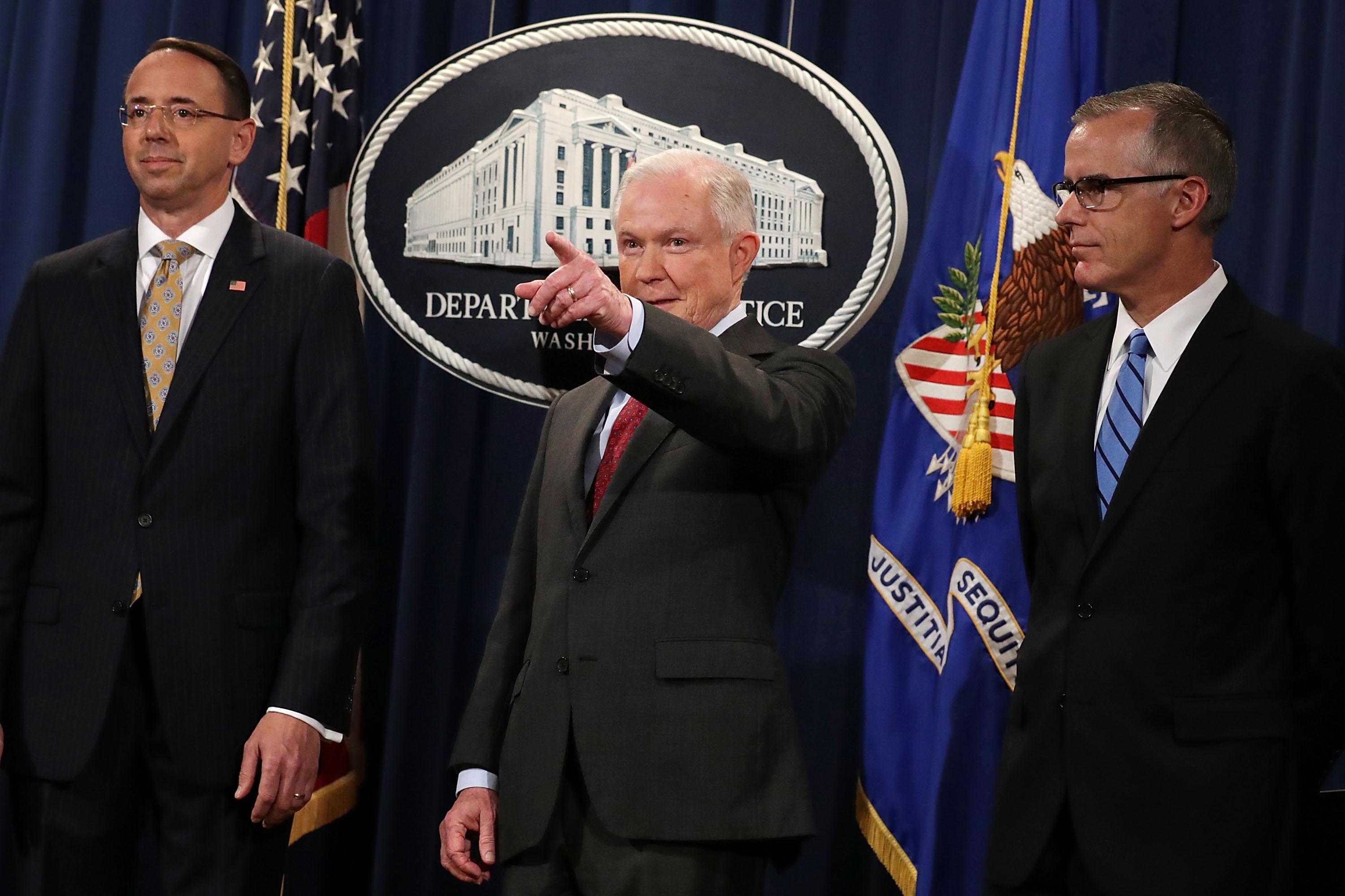 Rod Rosenstein, Jeff Sessions, and Andrew McCabe stand in front of a DOJ backdrop. Sessions points toward the assembled crowd.