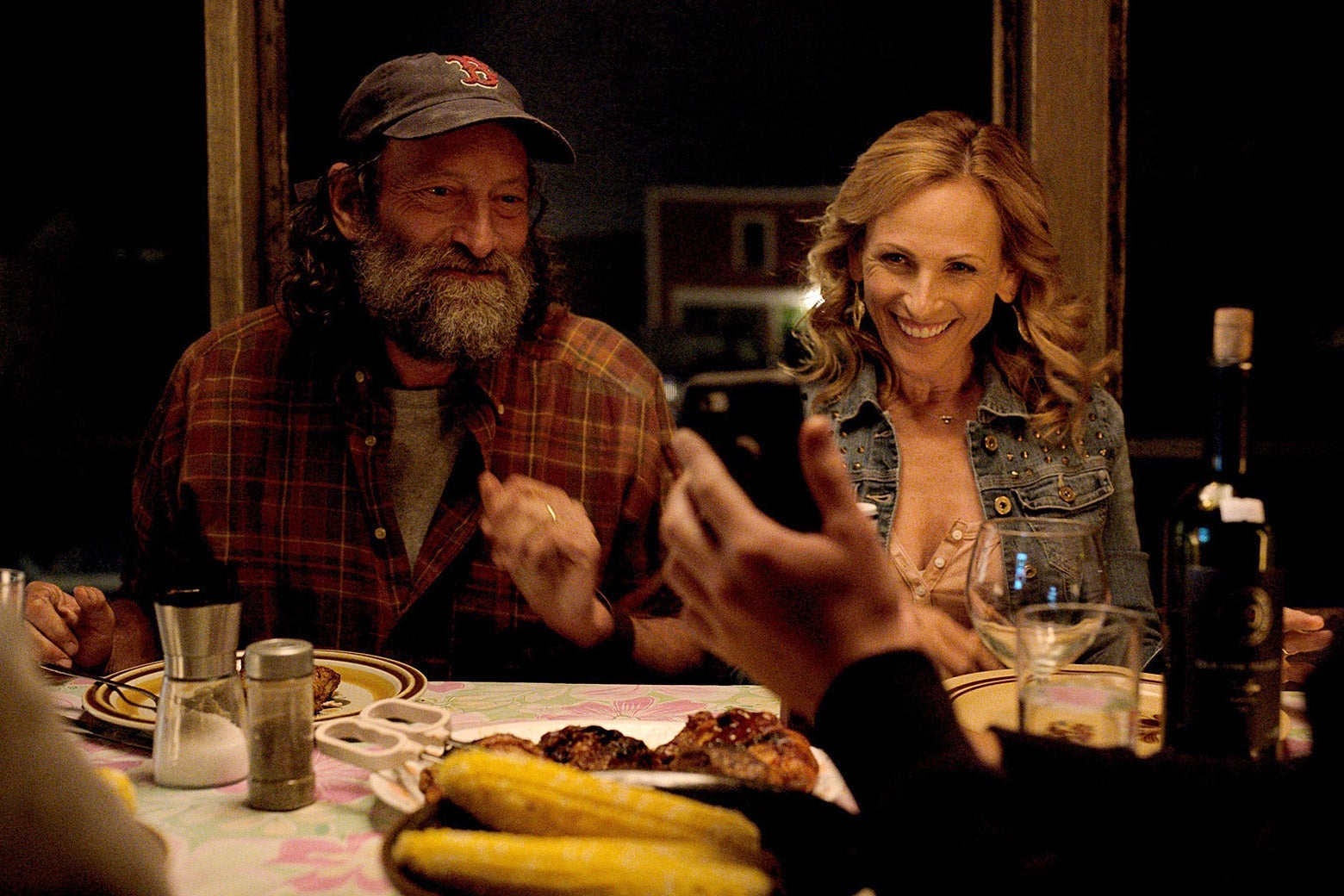 Troy Kotsur and Marlee Matlin, as Frank and Jackie Rossi, smile across a dinner table at a smartphone held by an unseen figure's hand. Frank has a bushy, graying beard and wears a cap with the Red Sox logo.
