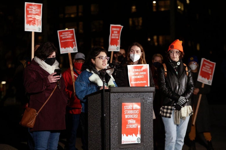 Four women wearing winter clothes stand at a lectern in front of a group holding red pro-union signs.