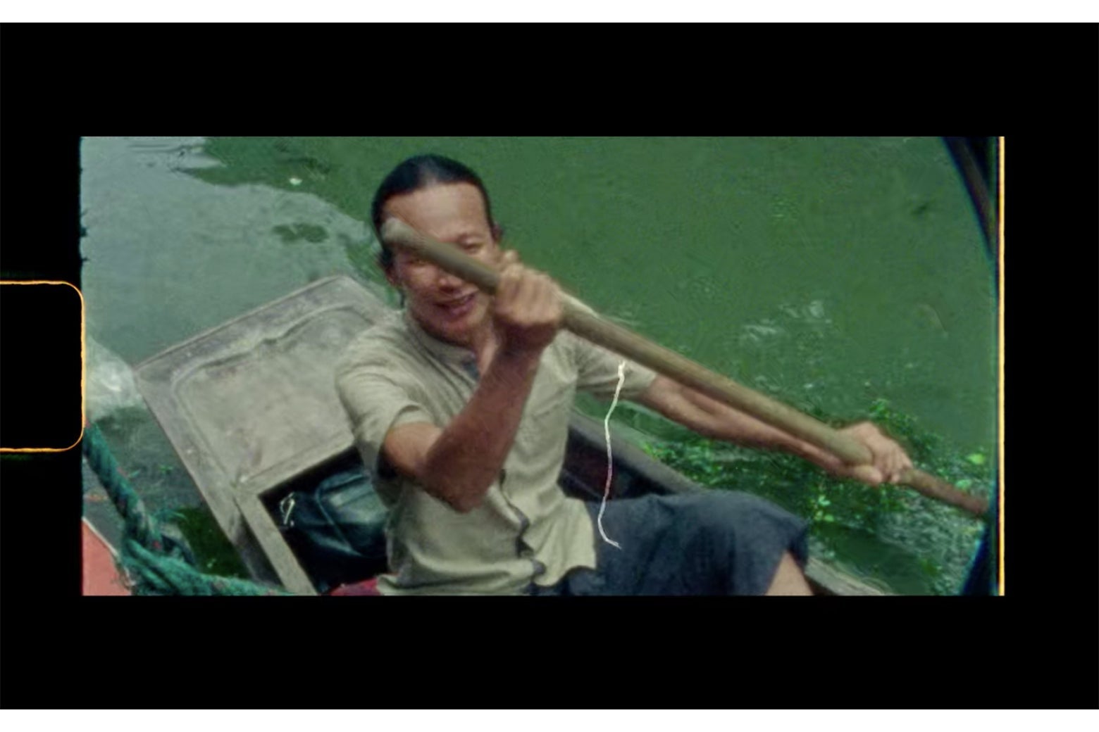 A man rows in a small boat on a river. A yellow curve is seen to the left, and a vertical yellow bar is seen on the right.