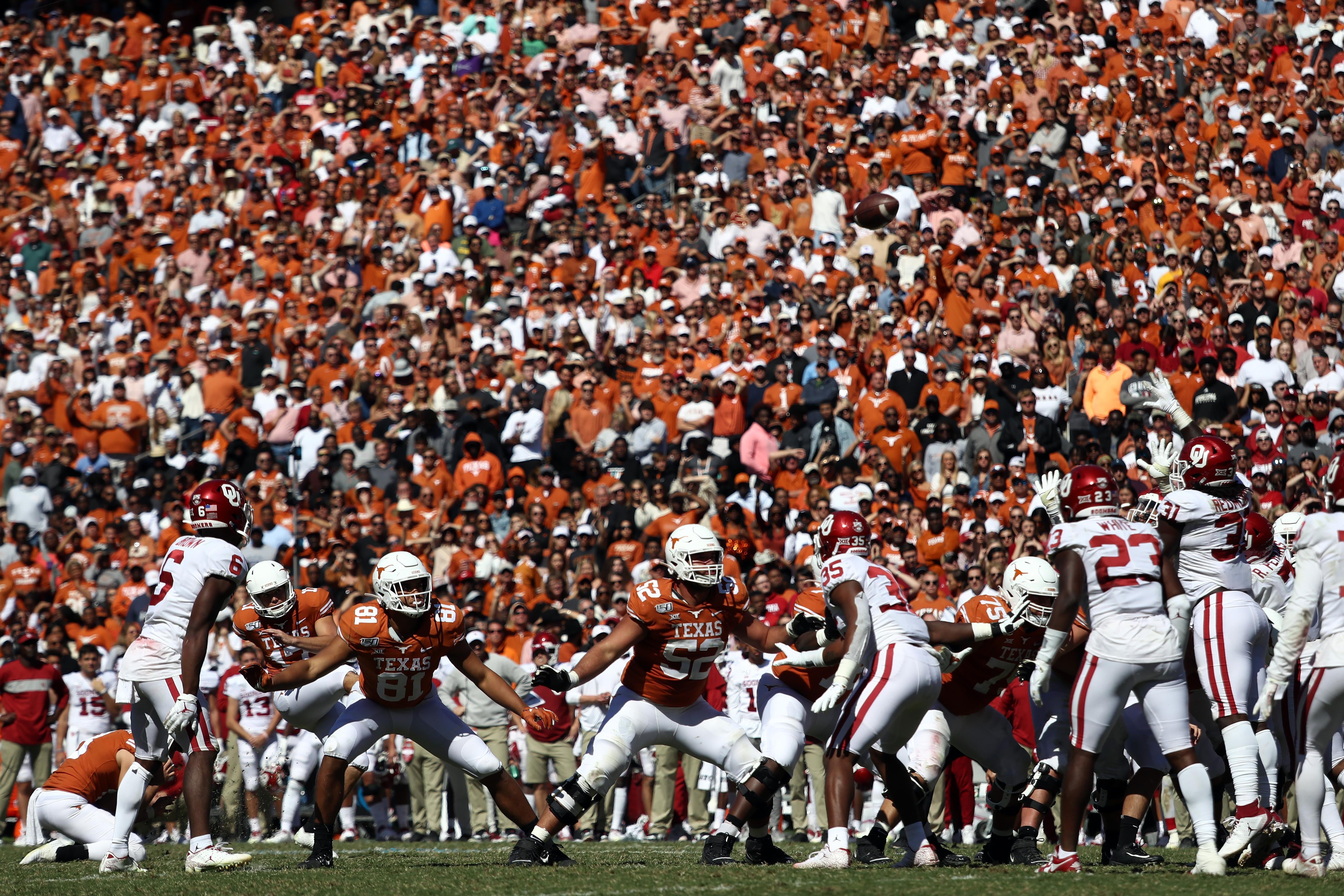 A scrum of Texas and Oklahoma players on the football field with the crowd in Texas orange and Oklahoma red looking on in the background