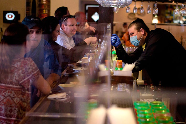A bartender in a face masks raises his hand to fist-bump a customer across from him who is also raising her fist.