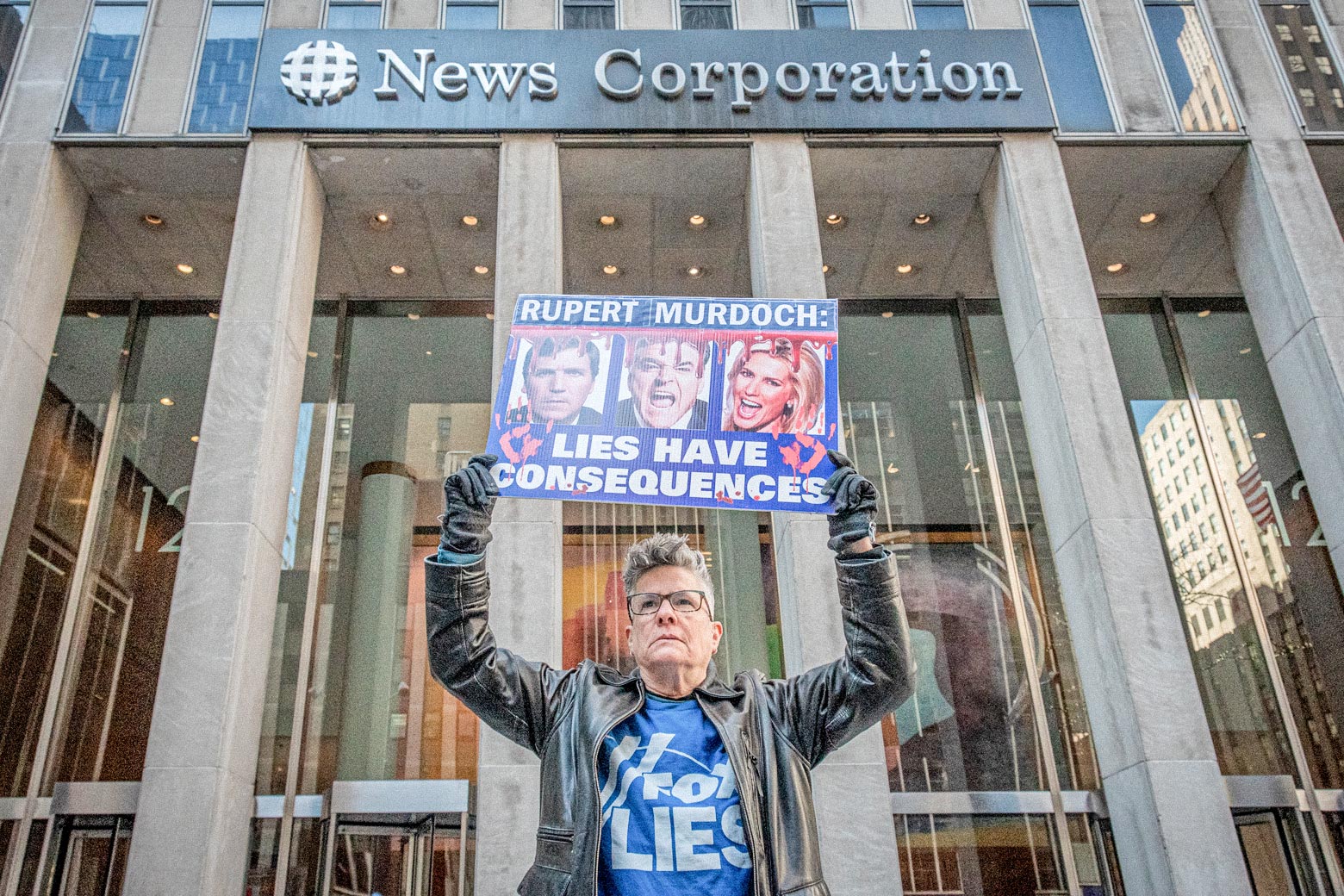 A woman holds a sign with the faces of Fox News hosts that says "Lies Have Consequences." She is standing in front of a skyscraper that is the headquarters of Fox News.