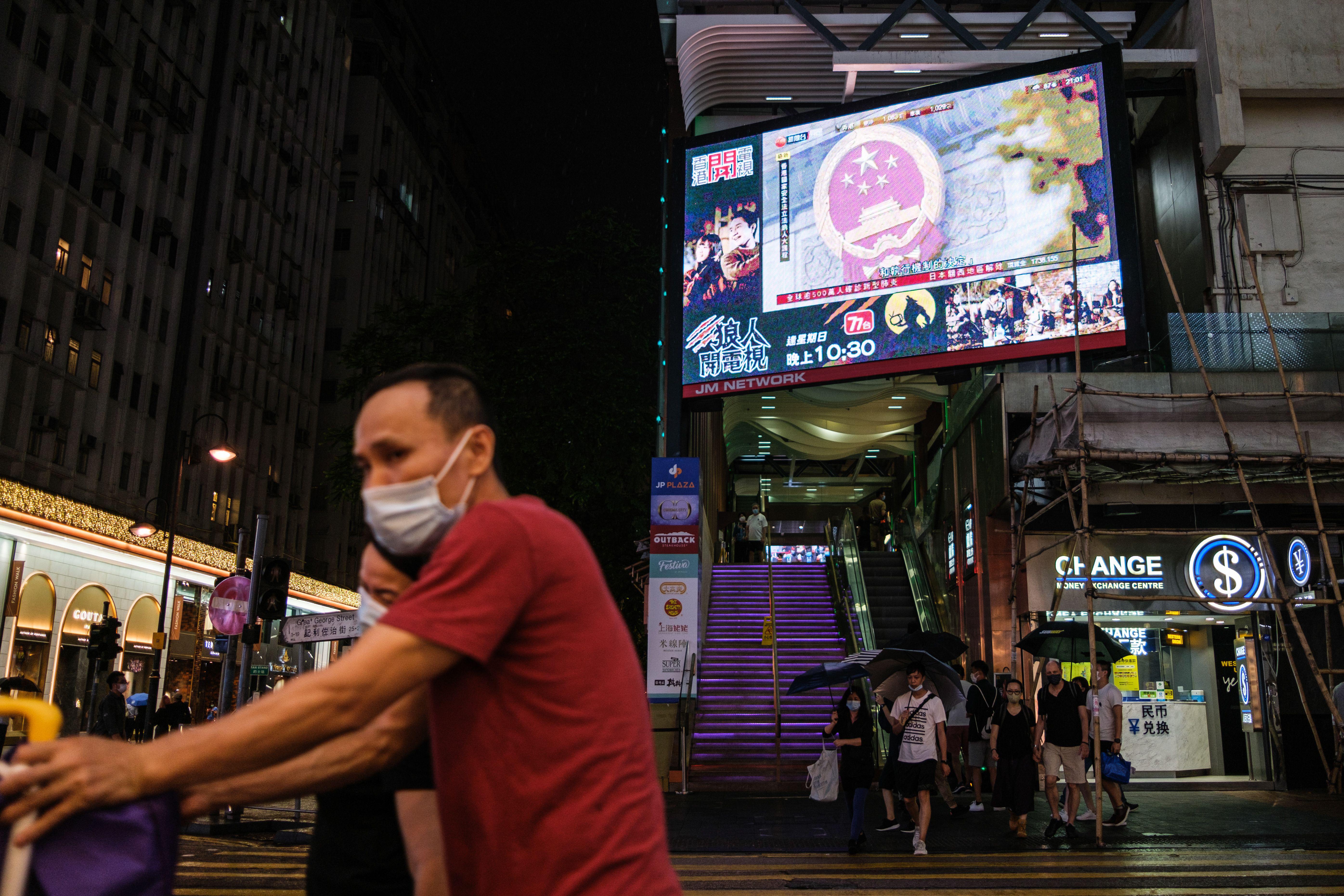 Pedestrians walk under a television screen in Hong Kong on May 21, 2020, showing a news broadcast from Beijing of the National Emblem of the People's Republic of China, ahead of the National People's Congress that opens on May 22. 