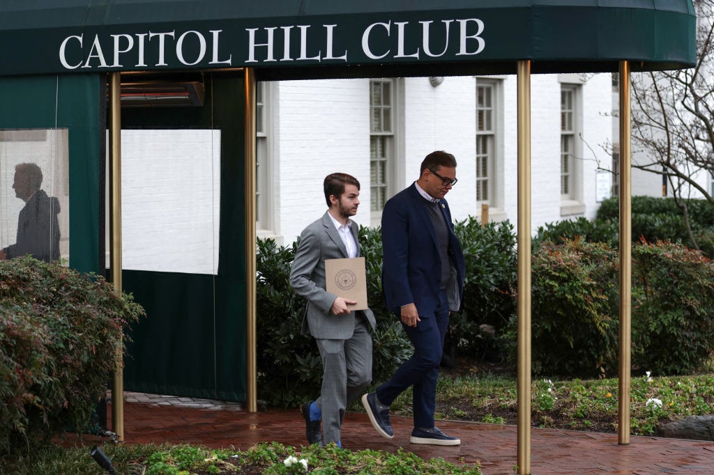 Santos, wearing a suit and sneakers, walks under an awning that reads "Capitol Hill Club" with a man who appears to be an aide.