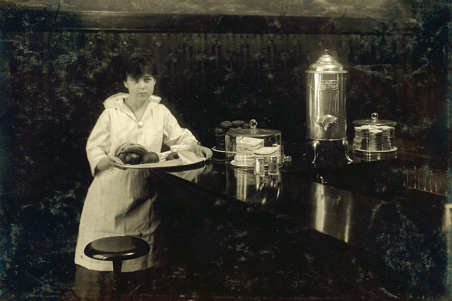 A young waitress wearing an apron and holding a tray leans on a restaurant counter next to cakes and a silver coffee pot.