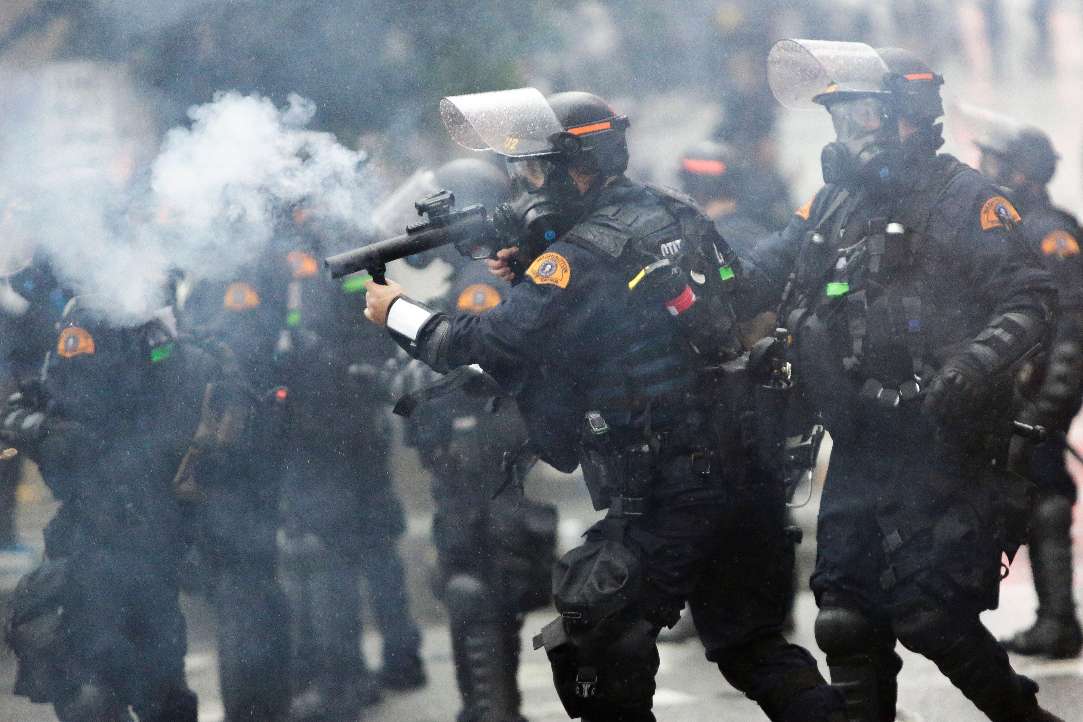 A row of officers in face shields, gas masks, and body armor firing tear gas canisters. 