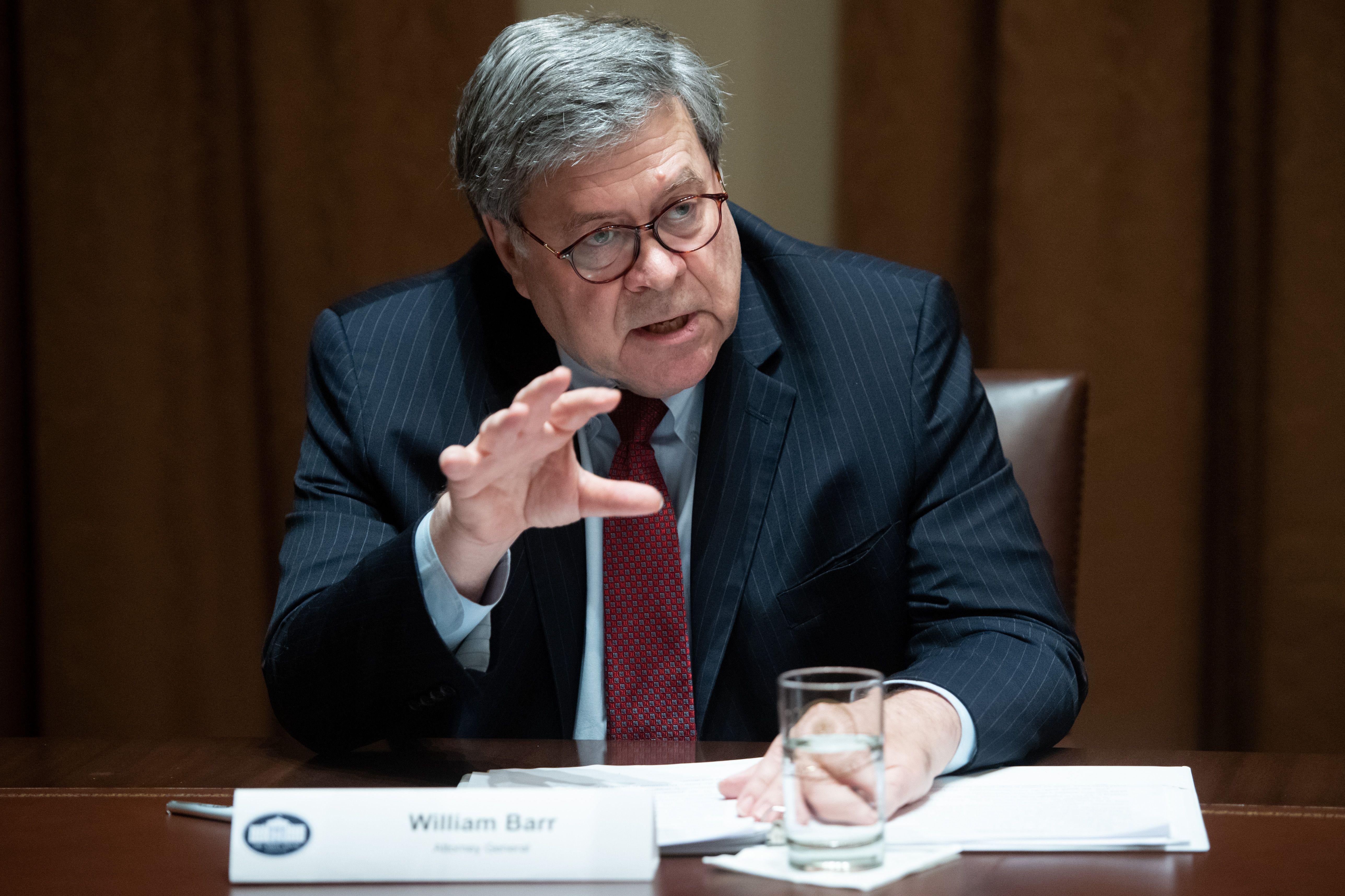 William Barr, sitting at a desk with a glass of water and some papers, raises his hand while speaking.