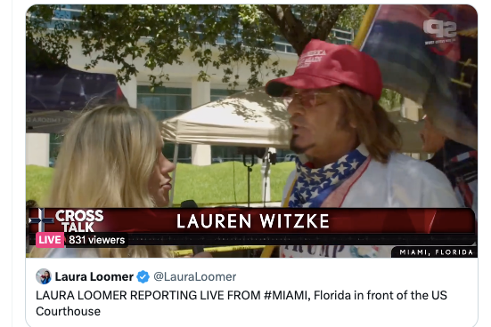 A blonde woman identified on screen as Lauren Witzke speaks to a man with a goatee and long, wispy hair who is wearing a Trump hat and kerchief and carrying a flag.