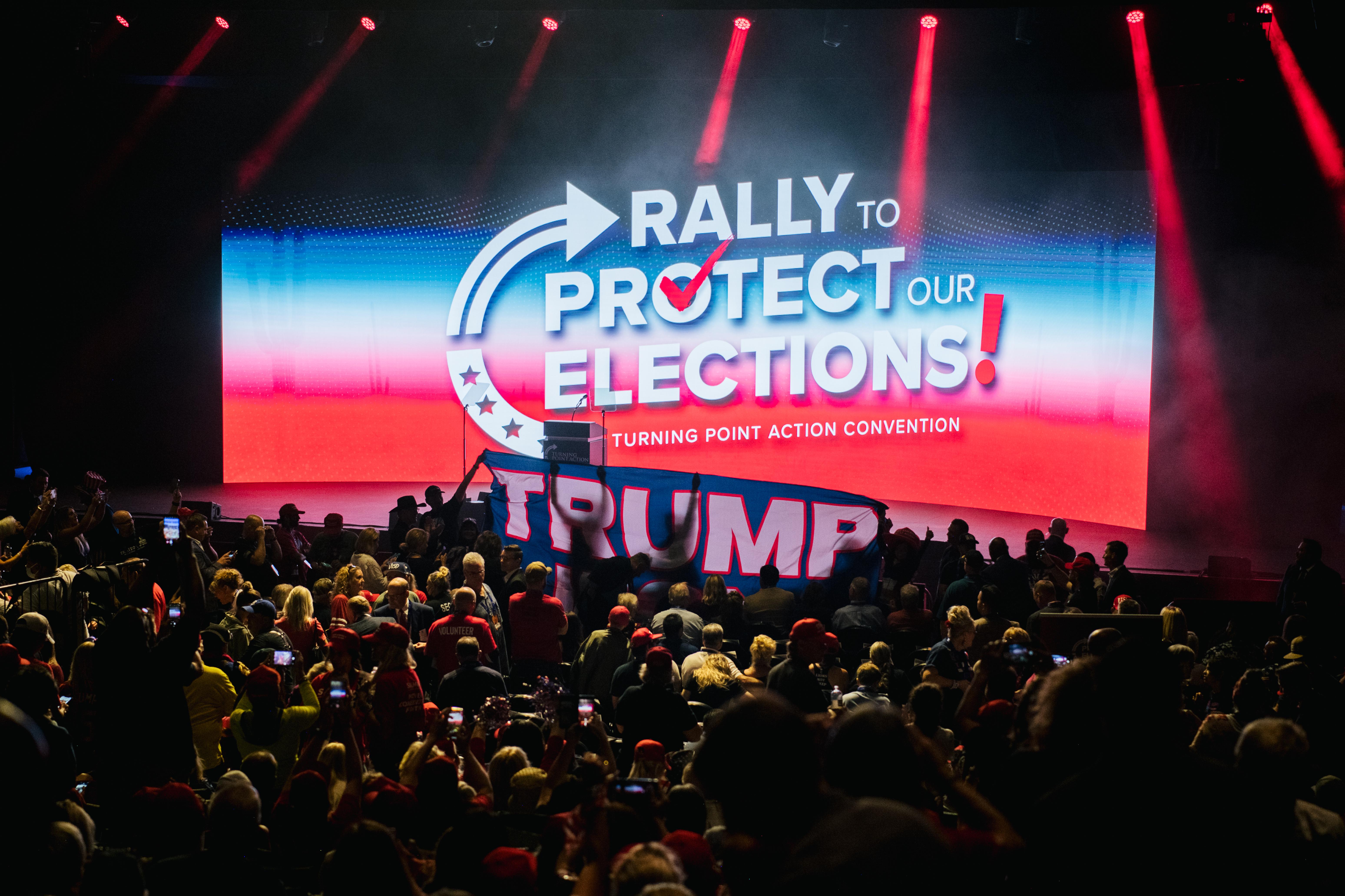 Many people seated in an auditorium indoors. On the stage is a Trump sign and a screen that says "Rally to Protect Our Elections Turning Point Action Convention."