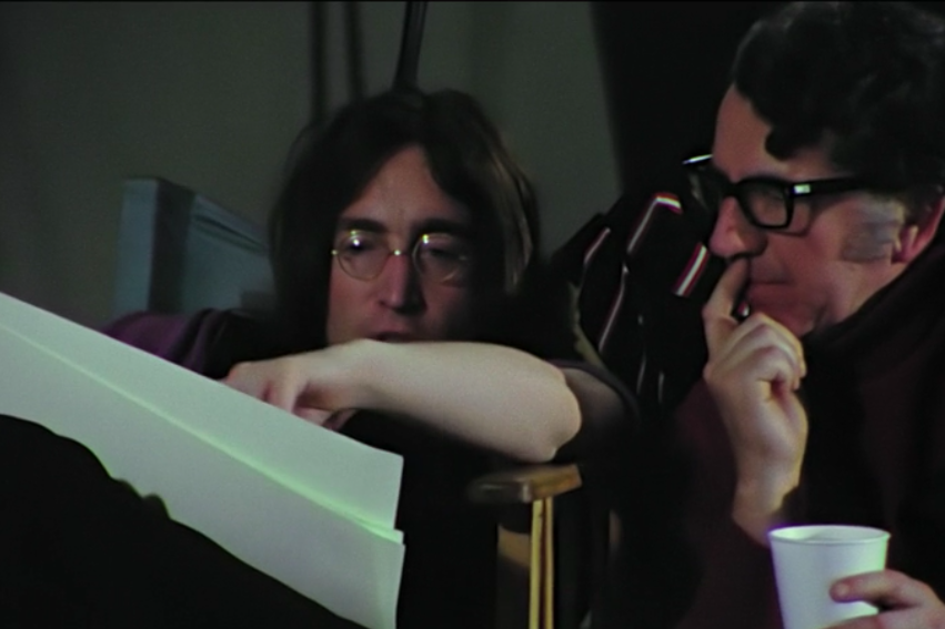 A man holding a disposable cup in one hand and putting his other hand to his mouth as he contemplates something on a piece of paper that John Lennon beside him is also looking at