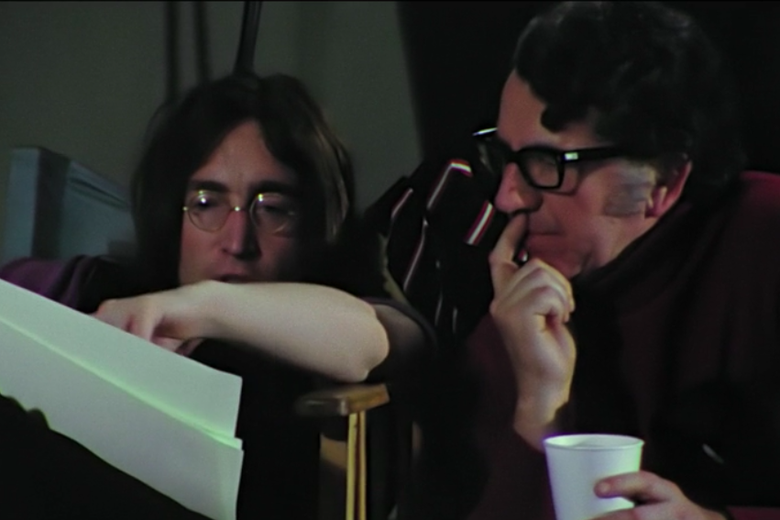 A man holding a disposable cup in one hand and putting his other hand to his mouth as he contemplates something on a piece of paper that John Lennon beside him is also looking at