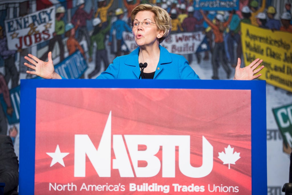 Warren, wearing a blue suit jacket, gestures with both hands from a lectern against a backdrop of drawings of workers wearing hard hats.