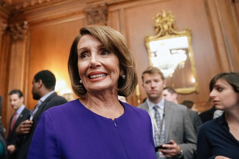 Nancy Pelosi wearing a blue jacket in a wood paneled room of the U.S. Capitol building surrounded by men in suits in the background.