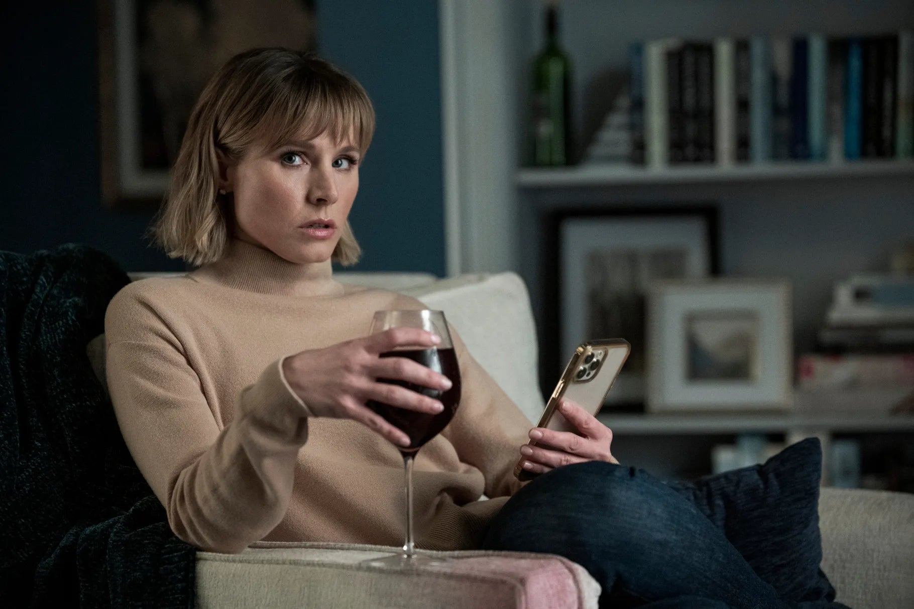 Bell sitting in an armchair with her phone in one hand and an overfull glass of red wine in the other hand as she looks to the side warily in a scene from the show