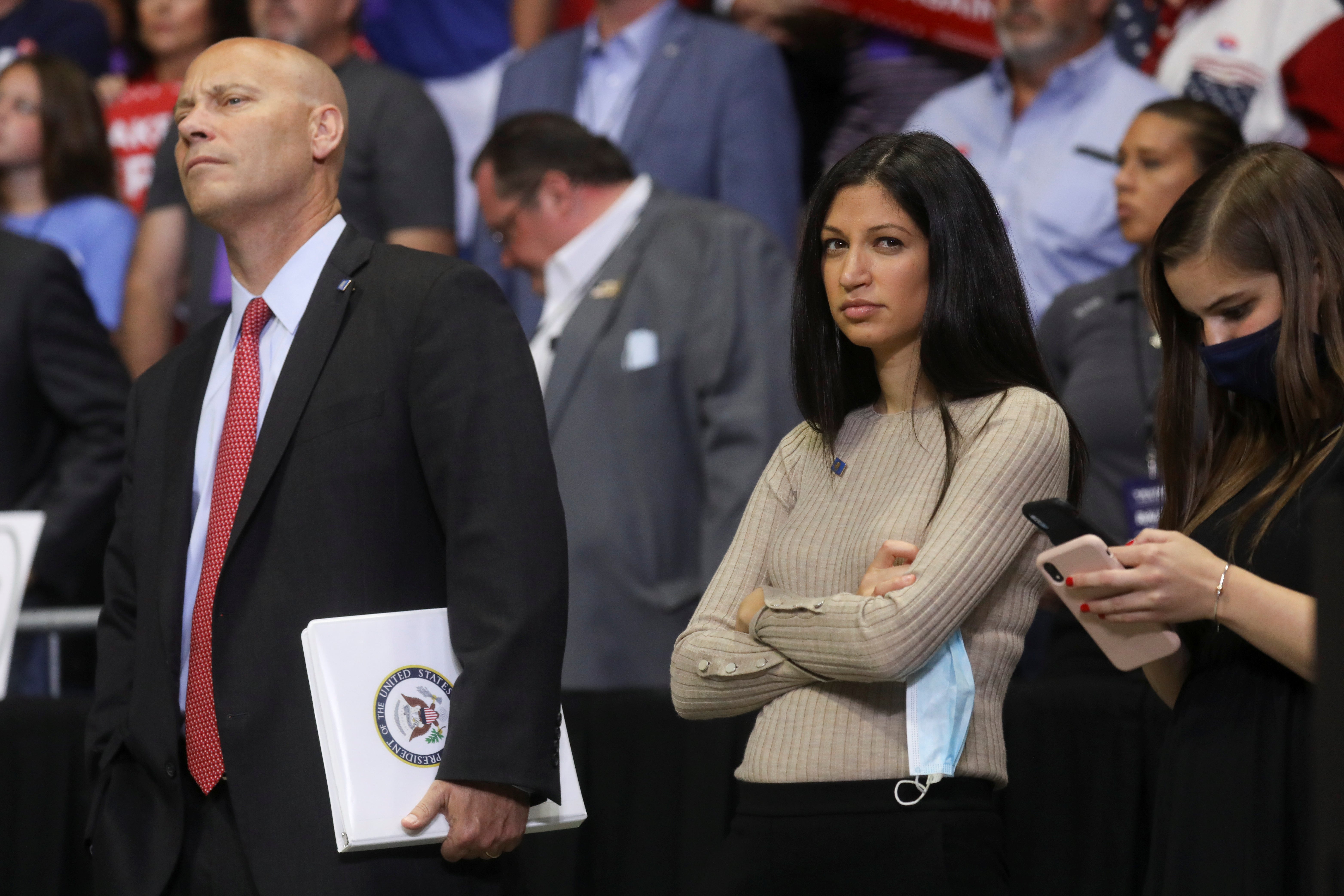 A white woman with long dark hair stands in an arena, arms crossed, looking skeptical.